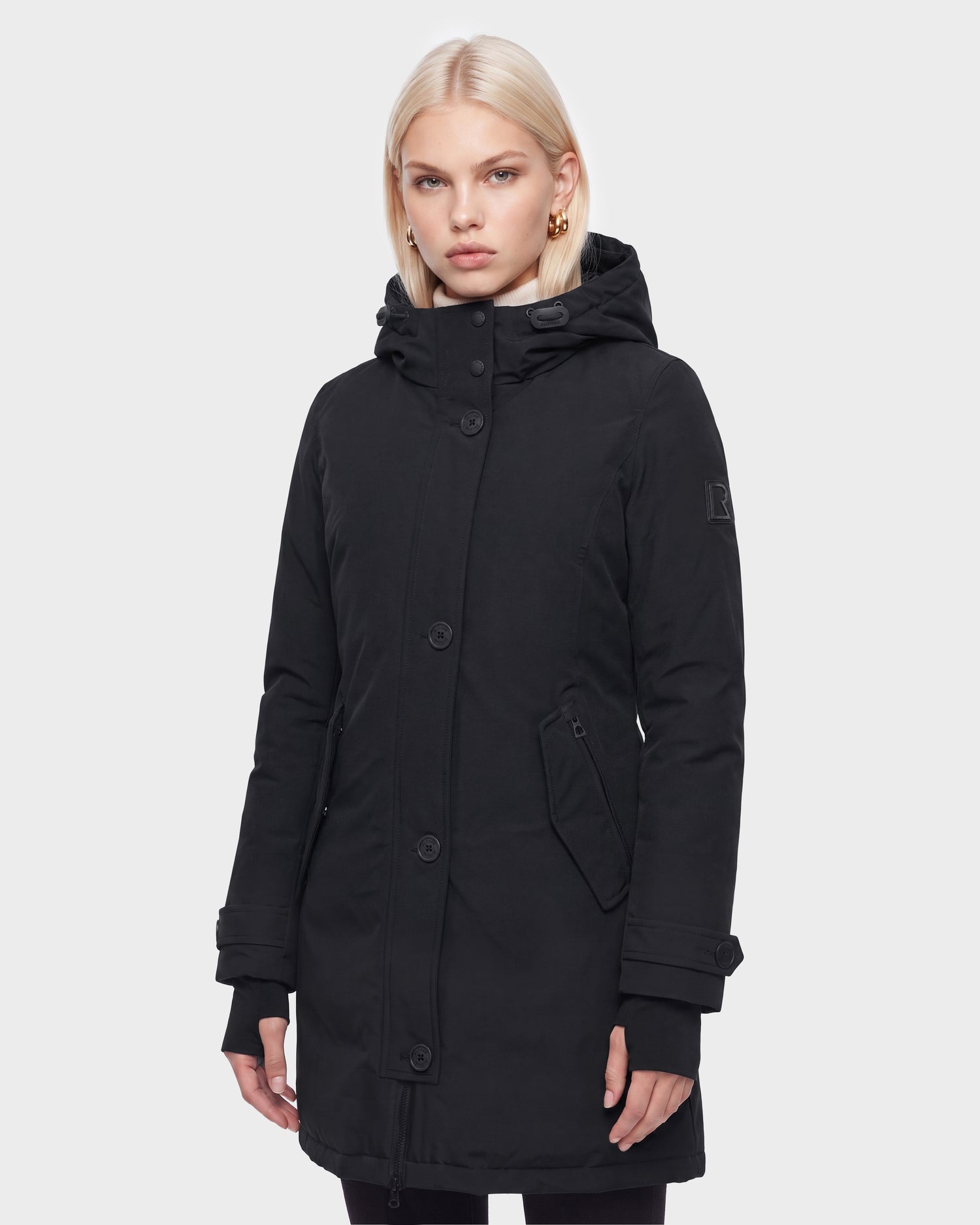 A woman with straight blonde hair wears the Rudsak KEYSHIA - 8126517 Black long hooded winter coat featuring buttons and zippered pockets, standing against a plain light gray background.