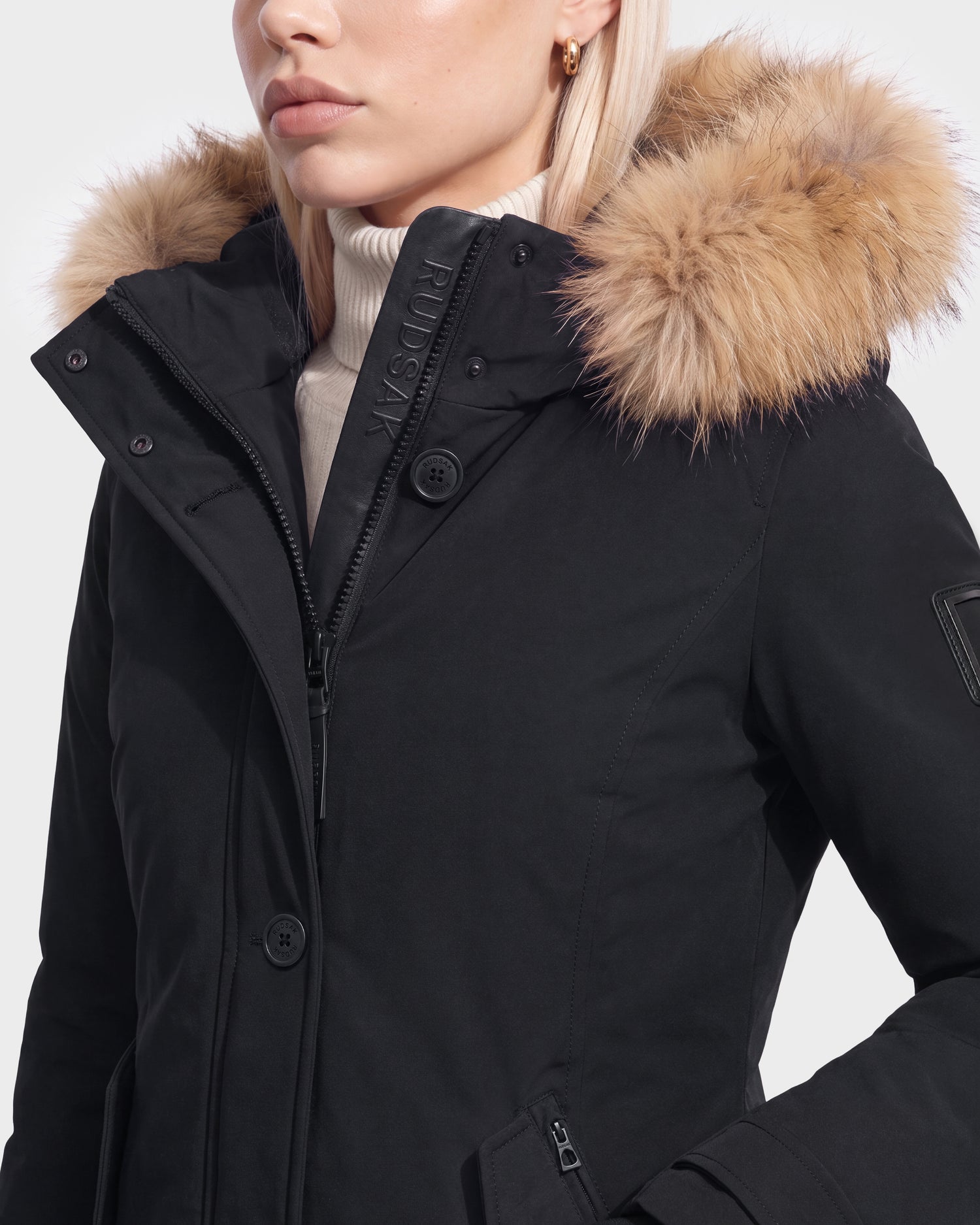 A person wearing the Rudsak KEYSHIA - 8126517 Black winter coat with a fur-lined hood and a white turtleneck sweater, shown from the shoulders up against a plain background.