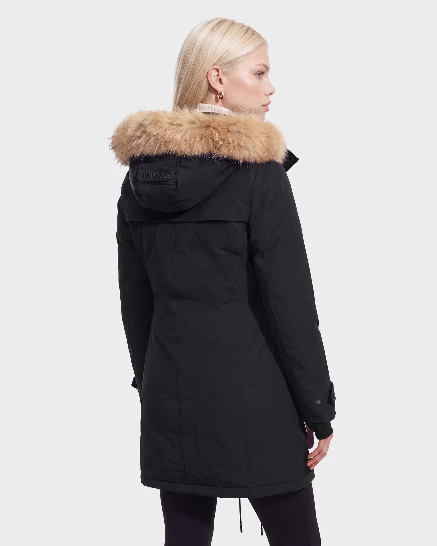 A woman with straight blonde hair stands with her back to the camera against a plain white background, wearing the Rudsak KEYSHIA - 8126517 Black parka featuring a fur-lined hood.