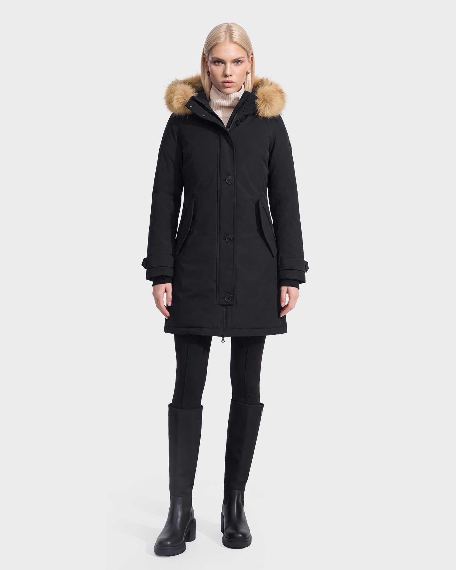 A woman wearing the Rudsak KEYSHIA - 8126517 Black parka with a fur-trimmed hood, paired with black leggings and black knee-high boots, stands against a plain white background.