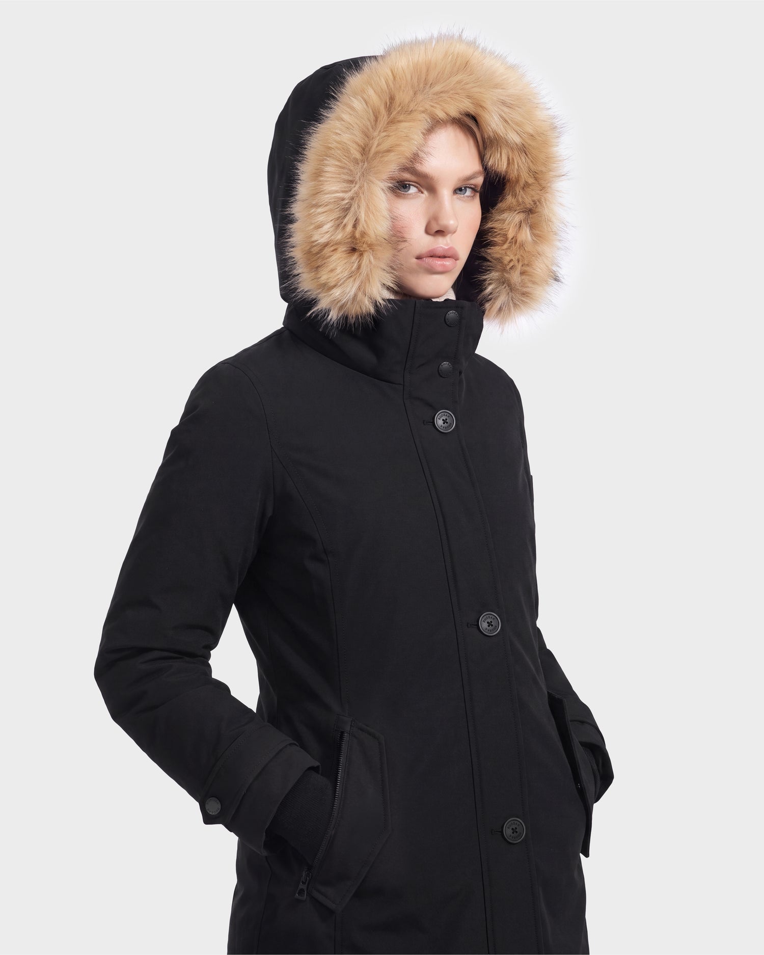 A person wearing the Rudsak KEYSHIA - 8126517 Black winter coat with a hood lined with tan faux fur, standing against a plain light background.