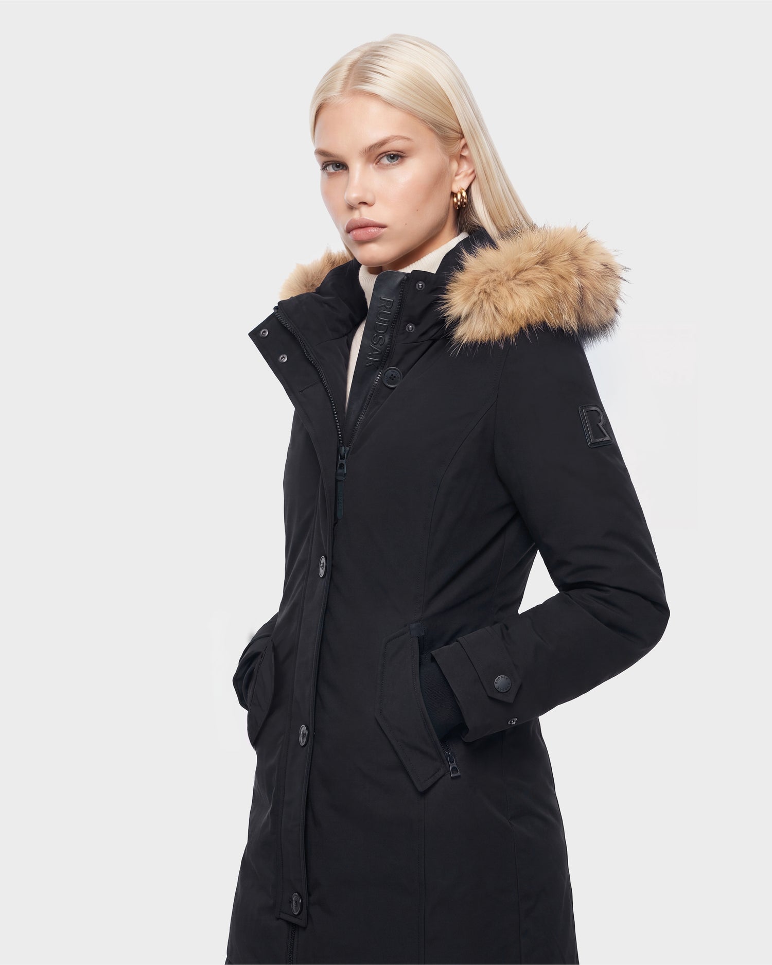 A woman with long blonde hair wears the Rudsak KEYSHIA - 8126517 Black coat, featuring a fur-trimmed hood. She stands with her hands in the pockets, facing slightly to the right against a plain light background.