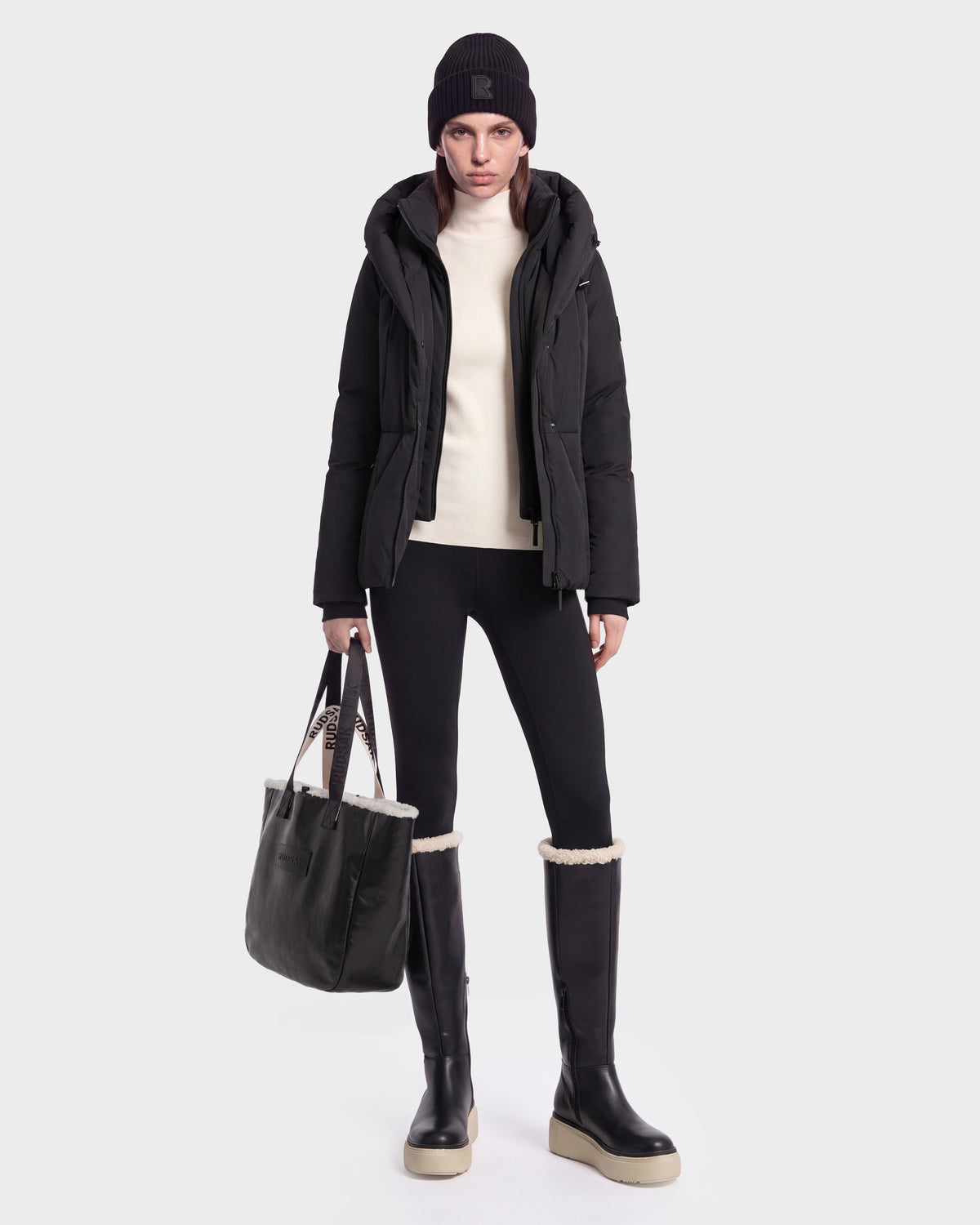 A woman wearing a short fitted puffer jacket with a hood, cream turtleneck, black leggings, and knee-high boots, holding a Rudsak KOLAD - 8126536 Black tote bag with branded straps.