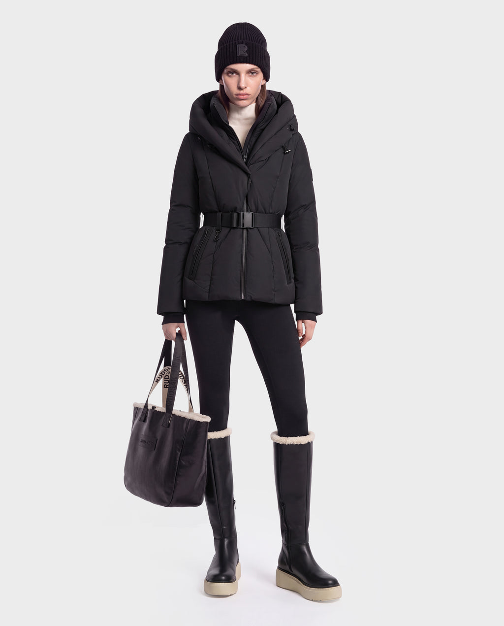 A model wears a black KOLAD - 8126536 short fitted puffer jacket, leggings, a beanie, and shearling-trimmed knee-high boots while holding a black tote bag.