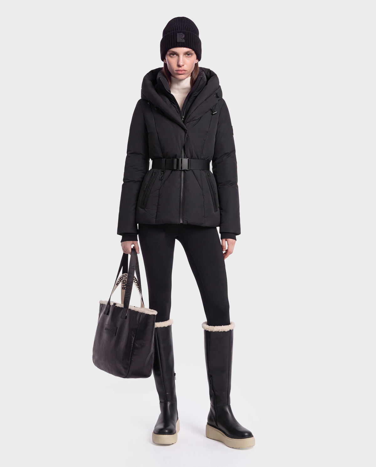 A model wears a black KOLAD - 8126536 short fitted puffer jacket, leggings, a beanie, and shearling-trimmed knee-high boots while holding a black tote bag.