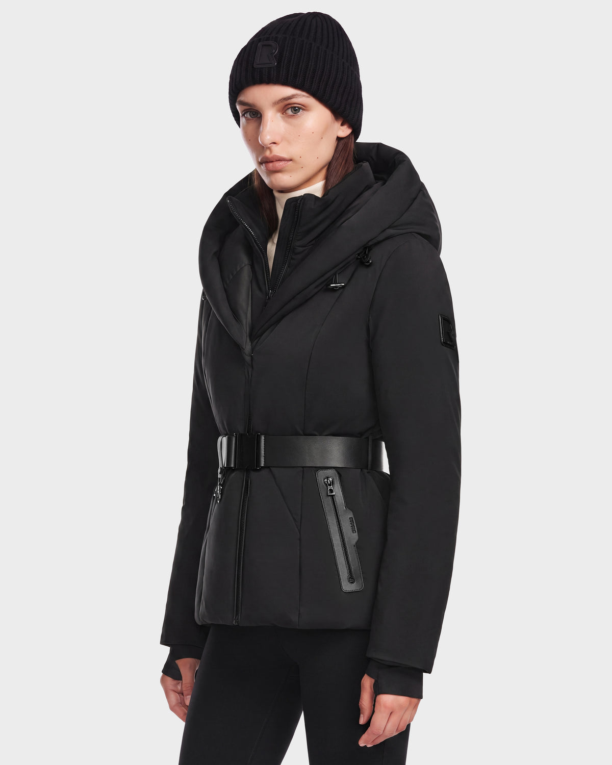 A person wearing a short fitted KOLAD - 8126536 Black puffer with a black hooded parka, wide buckled belt, and a matching knit beanie.