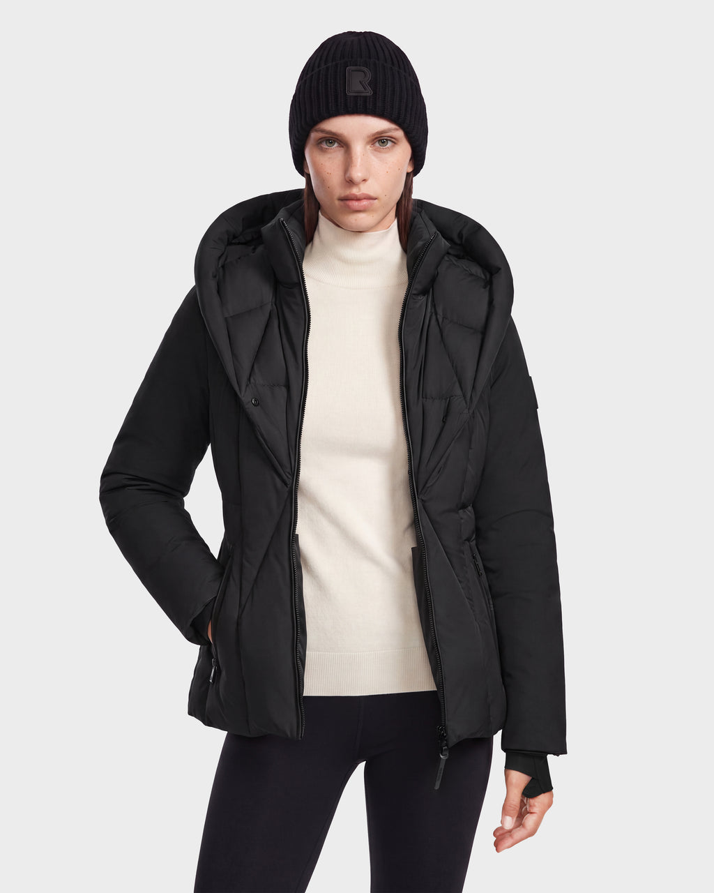 A person wearing a Rudsak KOLAD - 8126536 Black short fitted puffer jacket with a large hood, a white mock-neck sweater, and a black beanie.