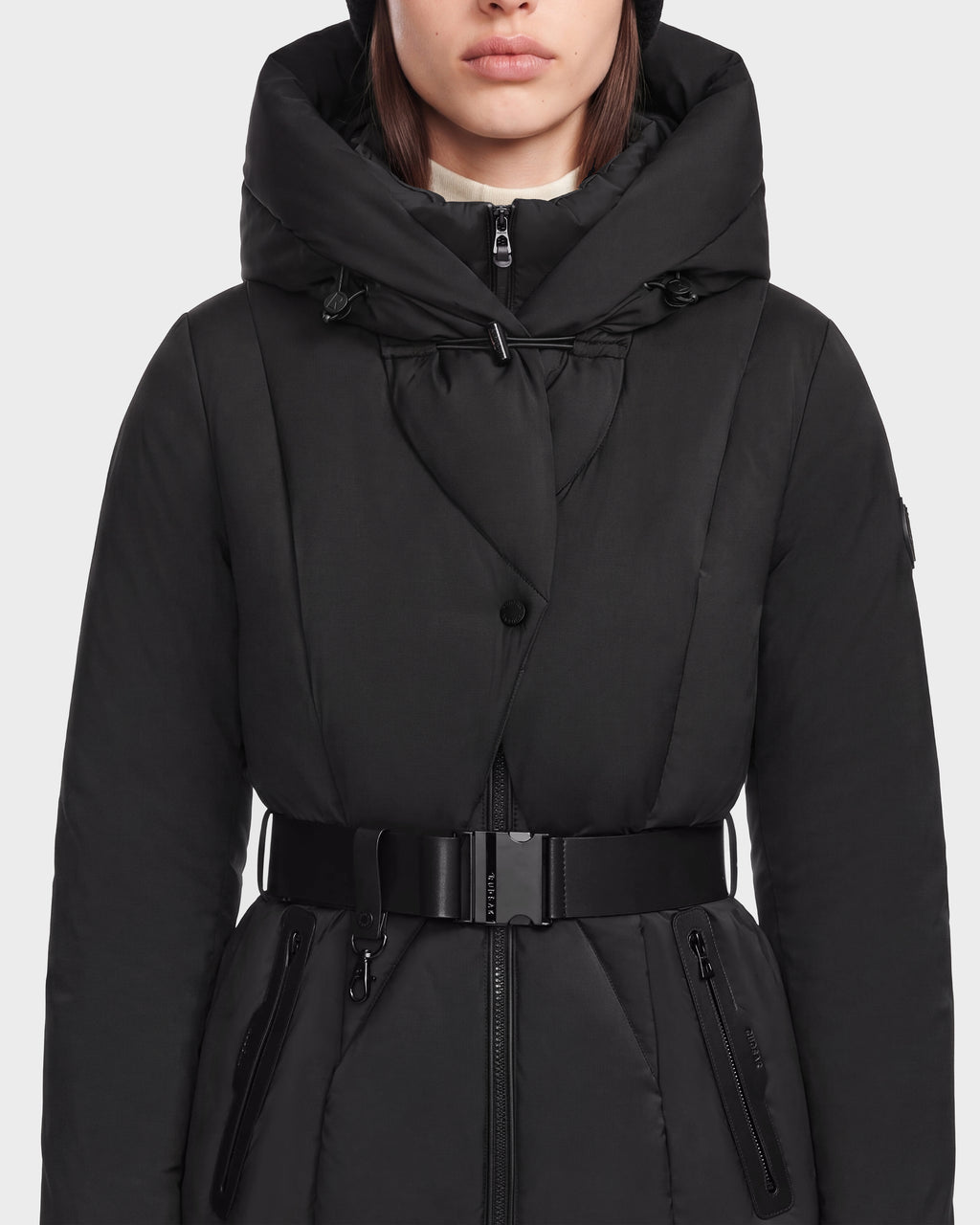 A woman stands against a plain white background wearing the Rudsak KOLAD - 8124528 Black puffer coat, which features RDS-certified down insulation, vegan leather trims, a large hood, and zippered pockets.