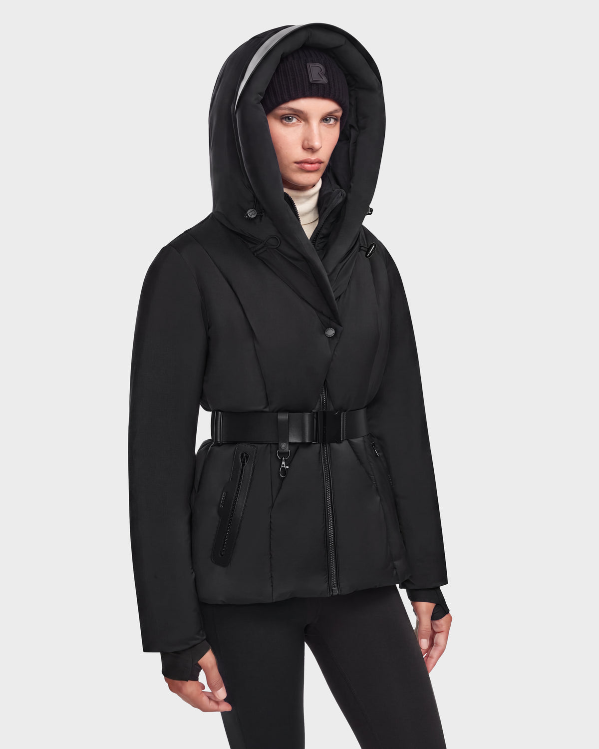 A person wearing the Rudsak KOLAD - 8124528 Black jacket, featuring RDS-certified down insulation and vegan leather trims, along with matching black pants and a black beanie, stands against a plain light background looking forward with a neutral expression.