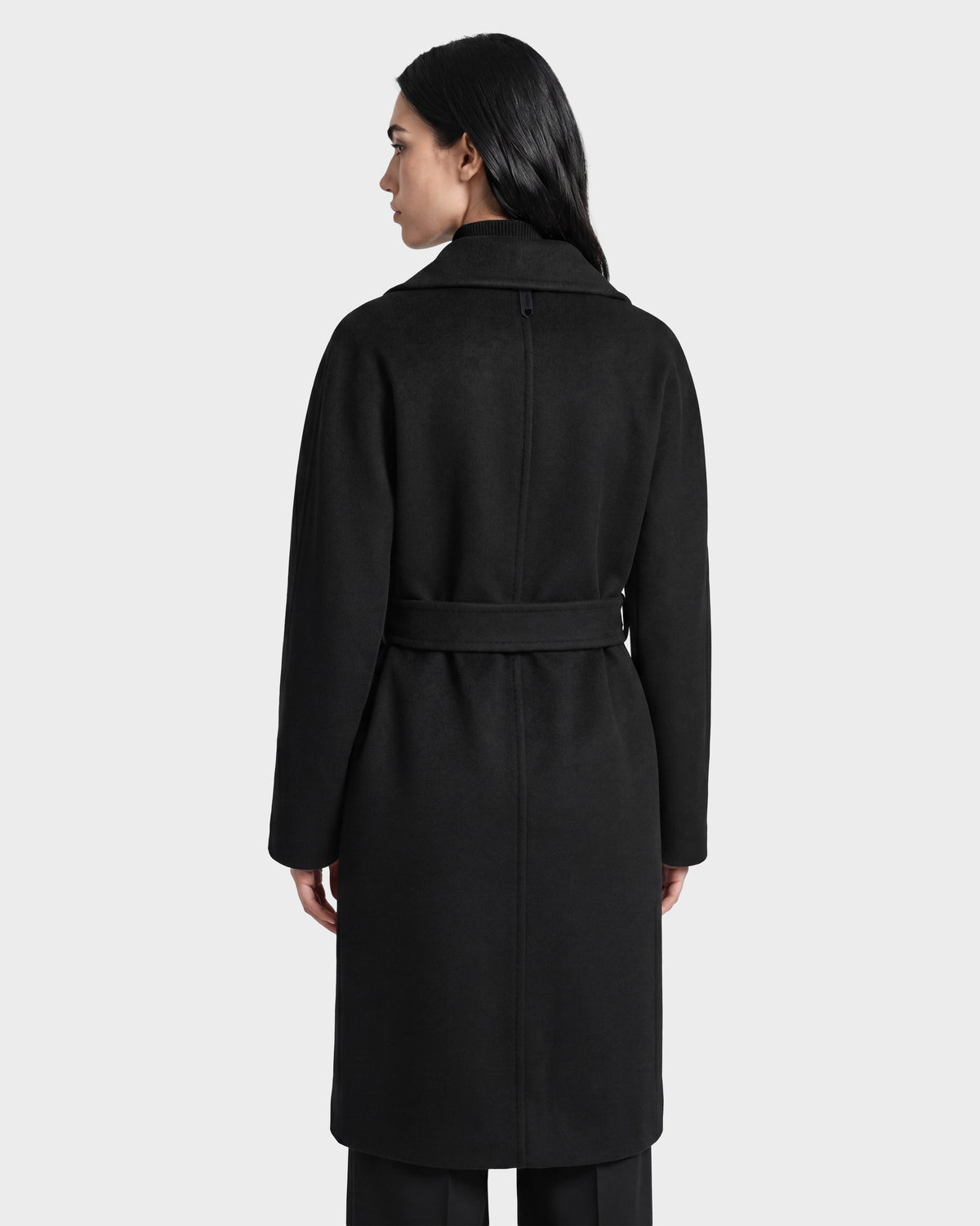 A woman with long dark hair, seen from behind, wears the Rudsak KOURT W - 8124702 Black belted wool coat, standing against a plain light gray background.