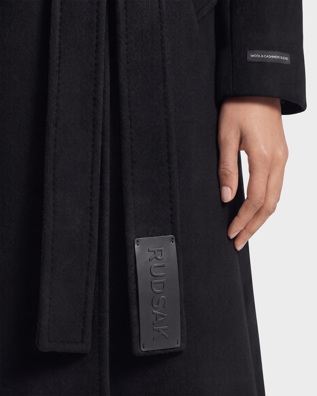 A close-up of a person wearing the KOURT W - 8124702 Black coat by Rudsak, featuring a belted design with "RUDSAK" on the belt and a sleeve tag reading "WOOL & CASHMERE BLEND." The person's hand rests relaxed by their side, highlighting the refined details of this elegant women's wool coat.