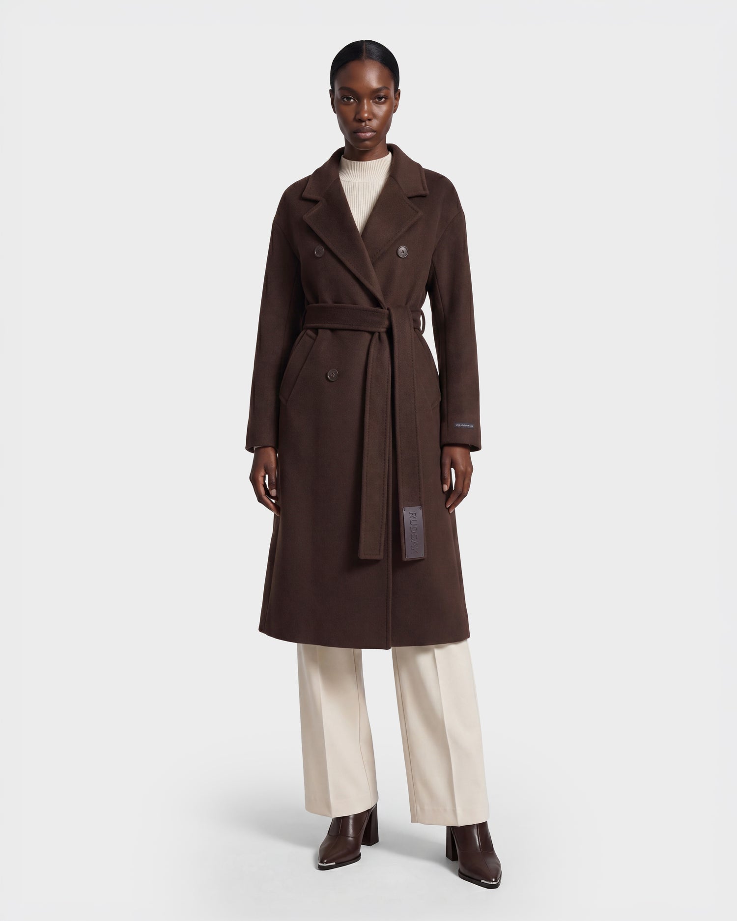 A person wearing the Rudsak KOURT W - 8124702 Espresso long, dark brown belted wool coat over a cream turtleneck and cream pants with brown ankle boots, standing against a plain white background.