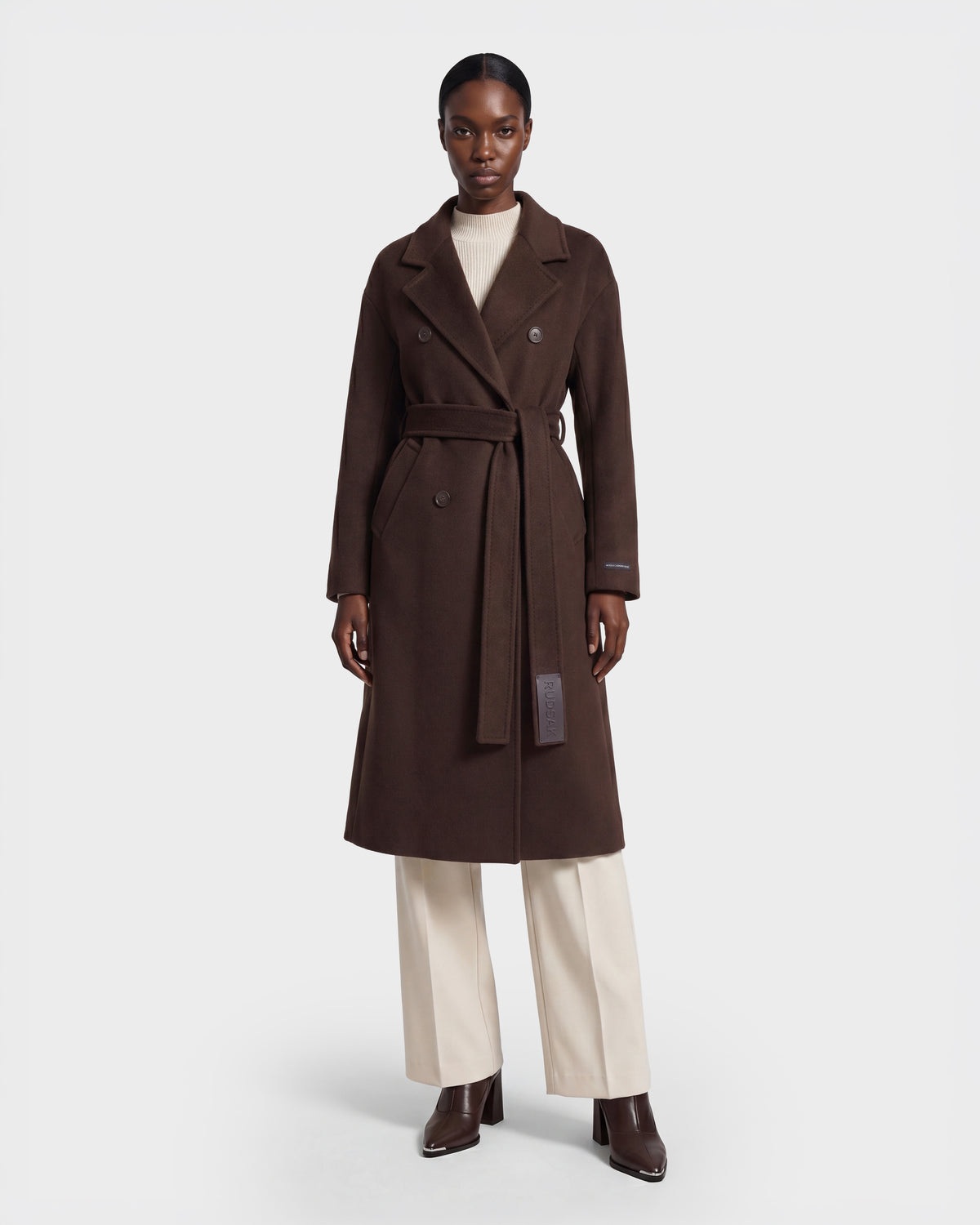 A person wearing the Rudsak KOURT W - 8124702 Espresso long, dark brown belted wool coat over a cream turtleneck and cream pants with brown ankle boots, standing against a plain white background.