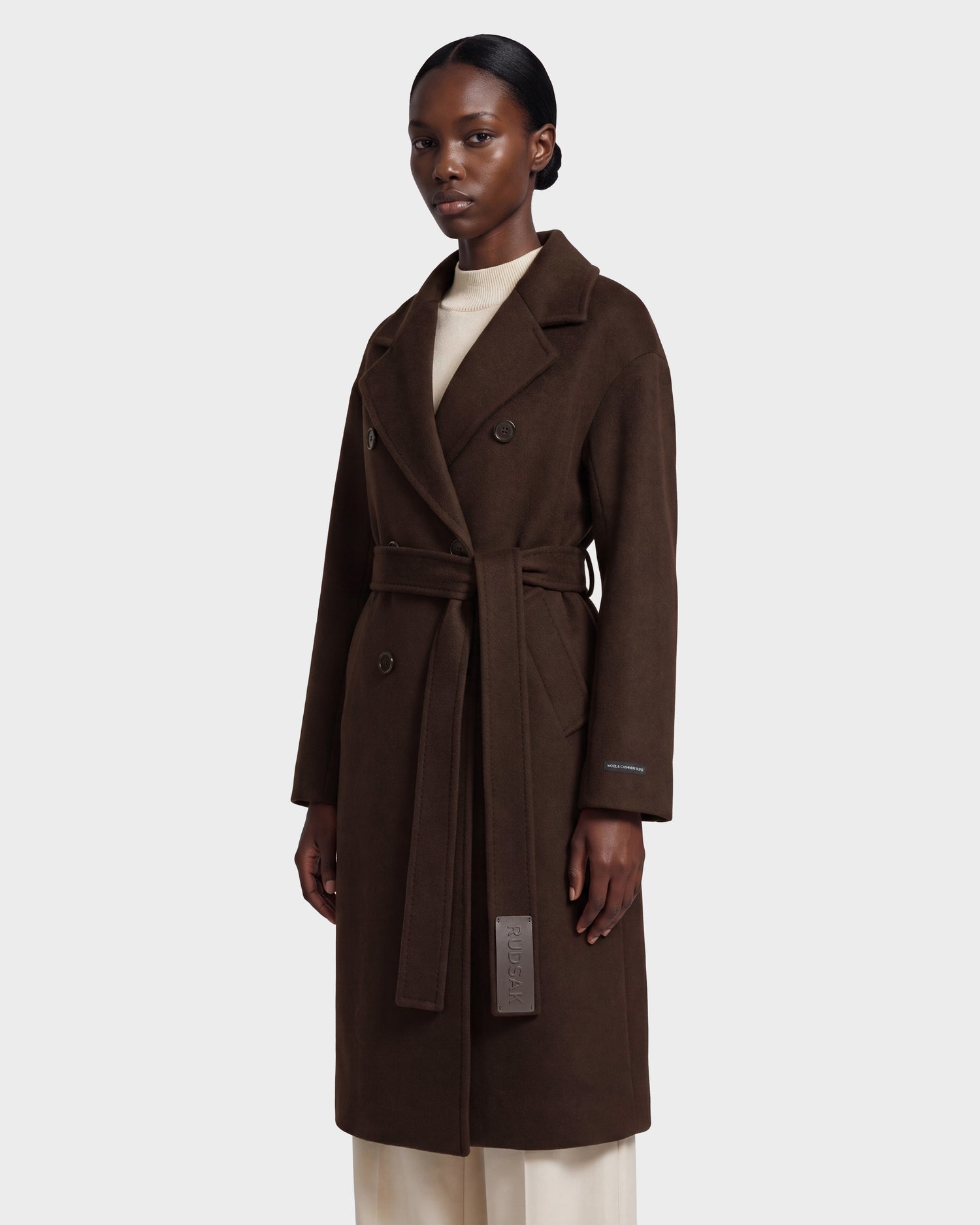 A woman wearing the Rudsak KOURT W - 8124702 Espresso, a long, dark brown double-breasted belted wool coat, over a light turtleneck and cream pants stands facing forward against a plain light background.