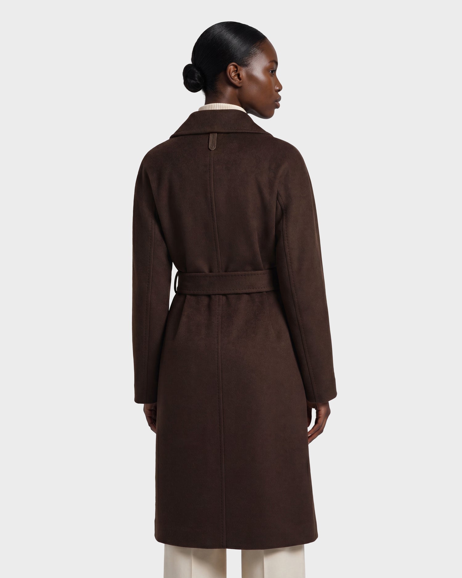 A woman with her hair in a low bun is seen from the back against a plain light background, wearing the Rudsak KOURT W - 8124702 Espresso, a long, dark brown belted wool coat, paired with light-colored pants.