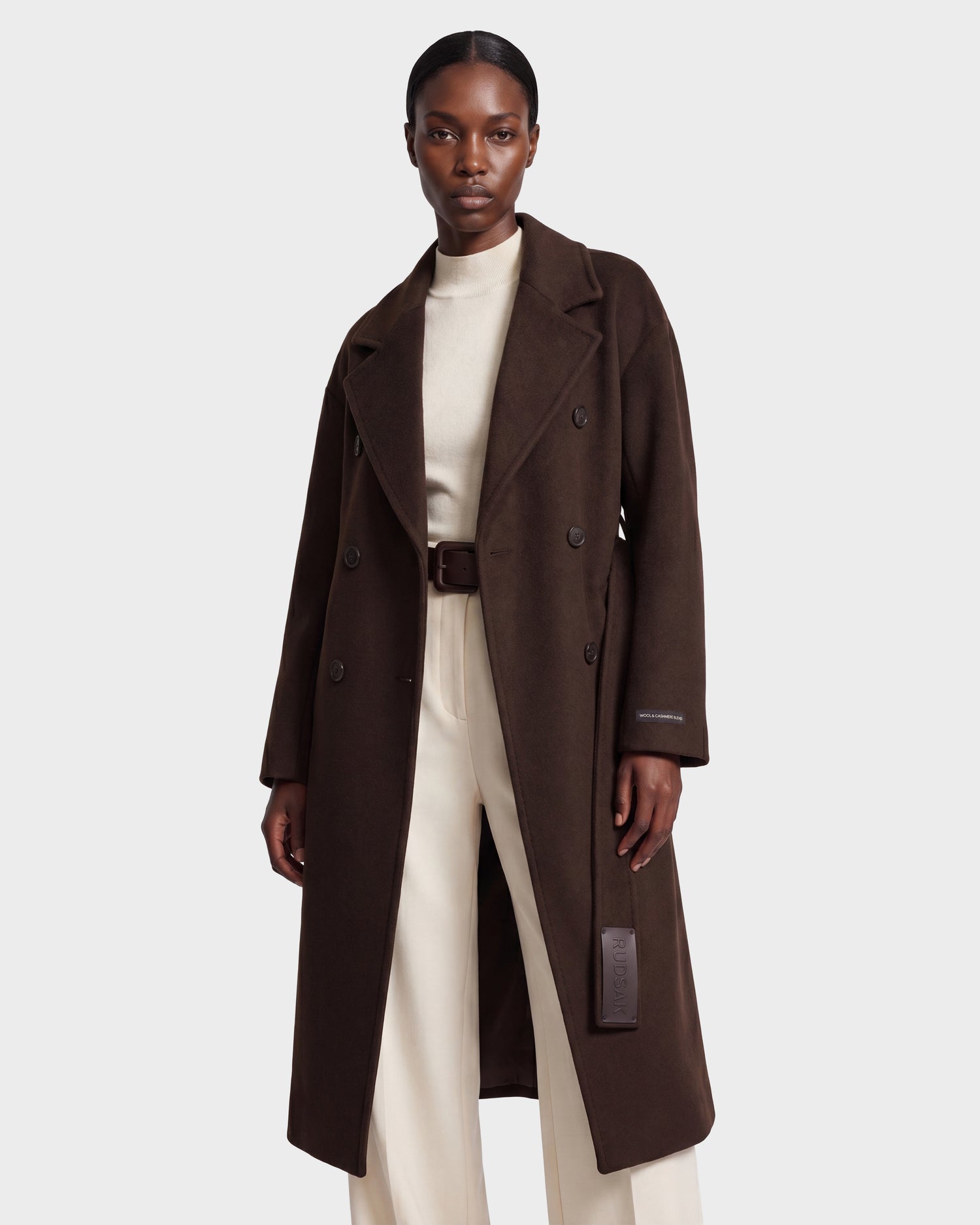 A woman wears the Rudsak KOURT W - 8124702 Espresso, a long, dark brown double-breasted women's wool coat, layered over a cream turtleneck and cream pants. She stands confidently against a plain light gray background.