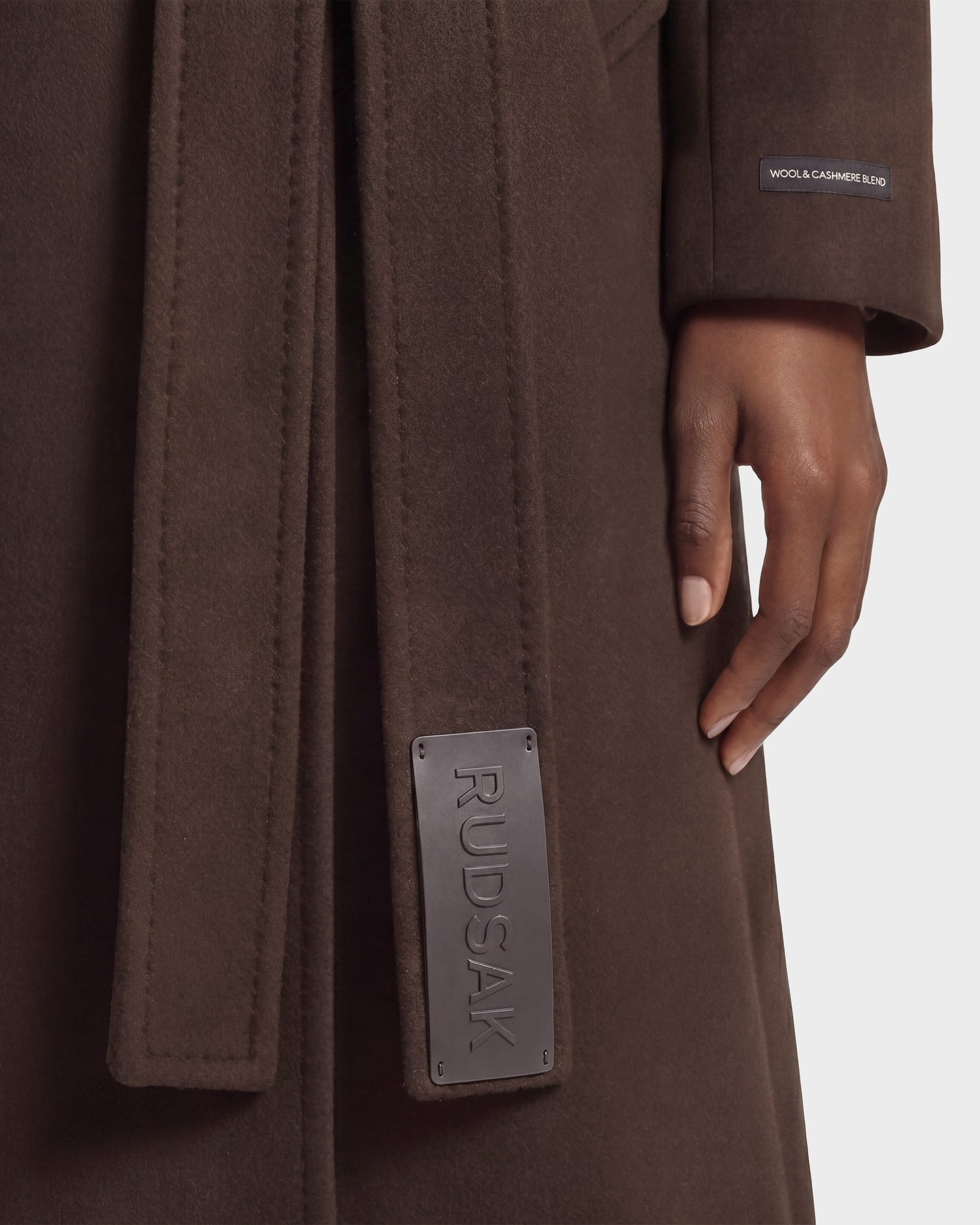 A person wearing the KOURT W - 8124702 Espresso coat by Rudsak—a brown wool and cashmere blend with a belted waist accented by a black leather “RUDSAK” tag; only the lower part of the arm and hand are visible.
