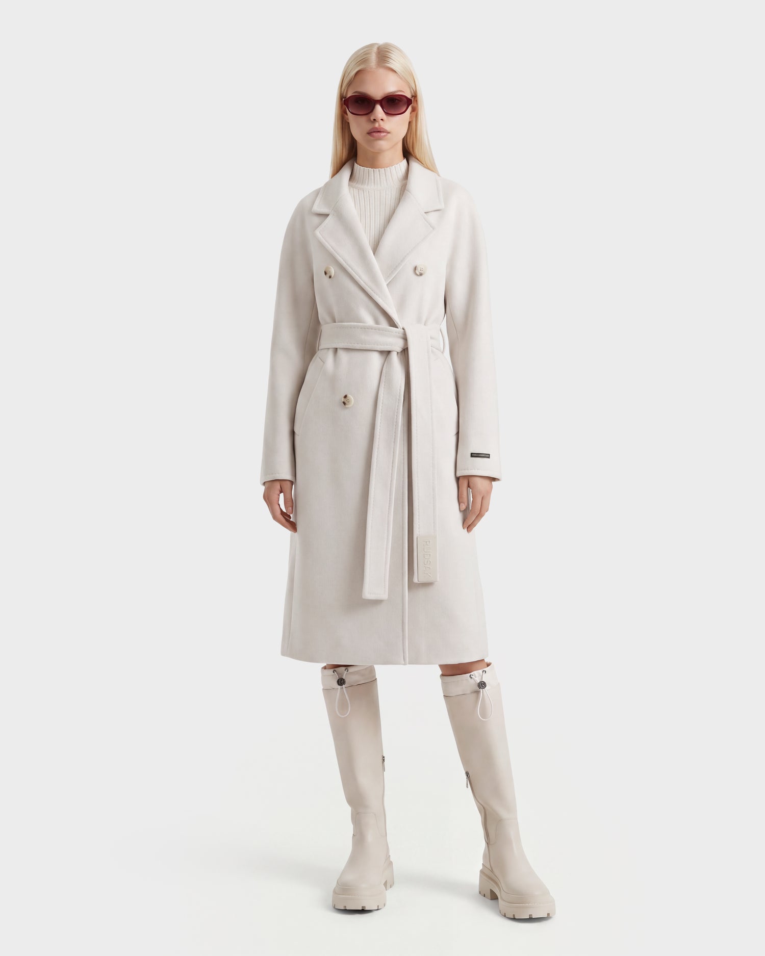 A woman wearing the Rudsak KOURT W - 8124702 Vanilla long belted wool coat, paired with a light turtleneck sweater, knee-high boots, and sunglasses, stands against a plain white background.