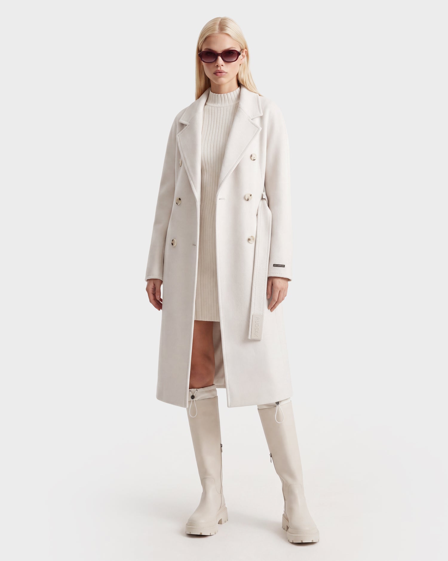 A person stands against a plain white background wearing the Rudsak KOURT W - 8124702 Vanilla, a luxurious long, white wool coat in a double-breasted style, paired with knee-high white boots, a white ribbed dress, and large sunglasses.