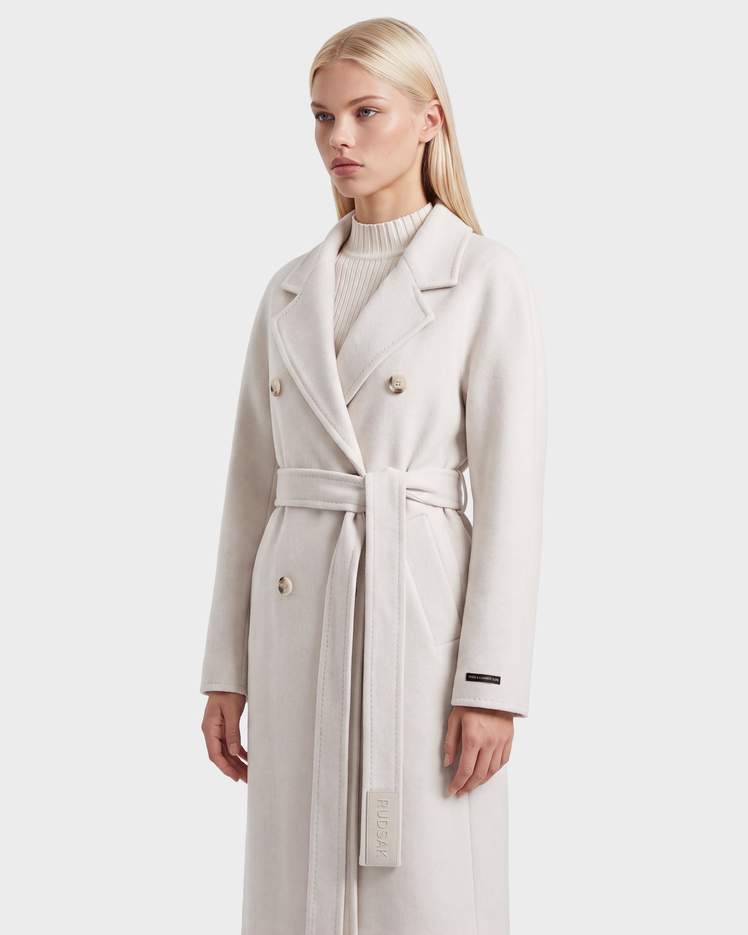 A woman with long blonde hair models the KOURT W - 8124702 Vanilla coat by Rudsak—a luxurious, light beige wool piece with a belted, double-breasted design—elegantly layered over a ribbed turtleneck sweater against a plain white background.