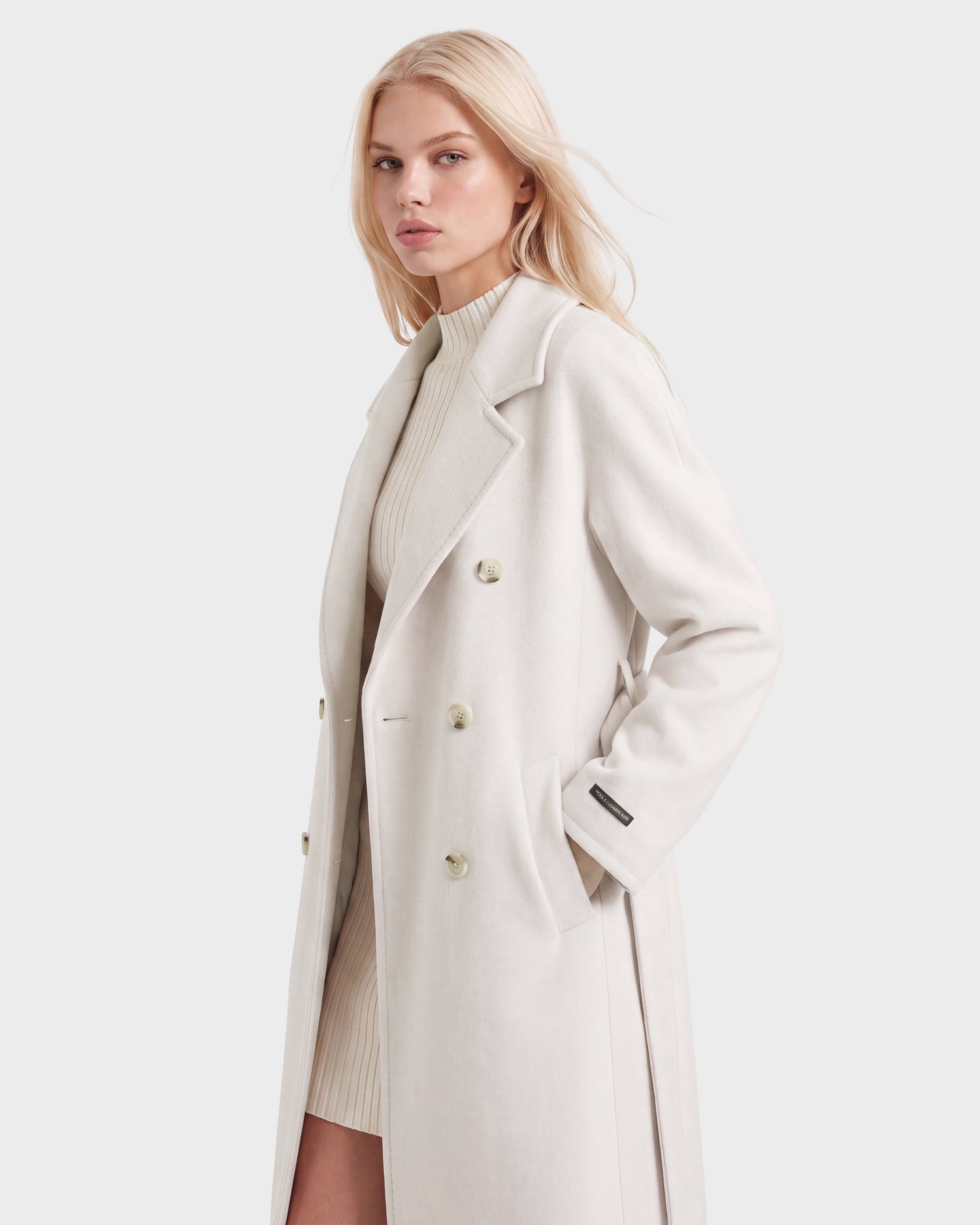 A woman with long blonde hair wears the KOURT W - 8124702 Vanilla, a luxurious double-breasted women's wool coat by Rudsak, over a ribbed light knit dress, standing against a plain light background.