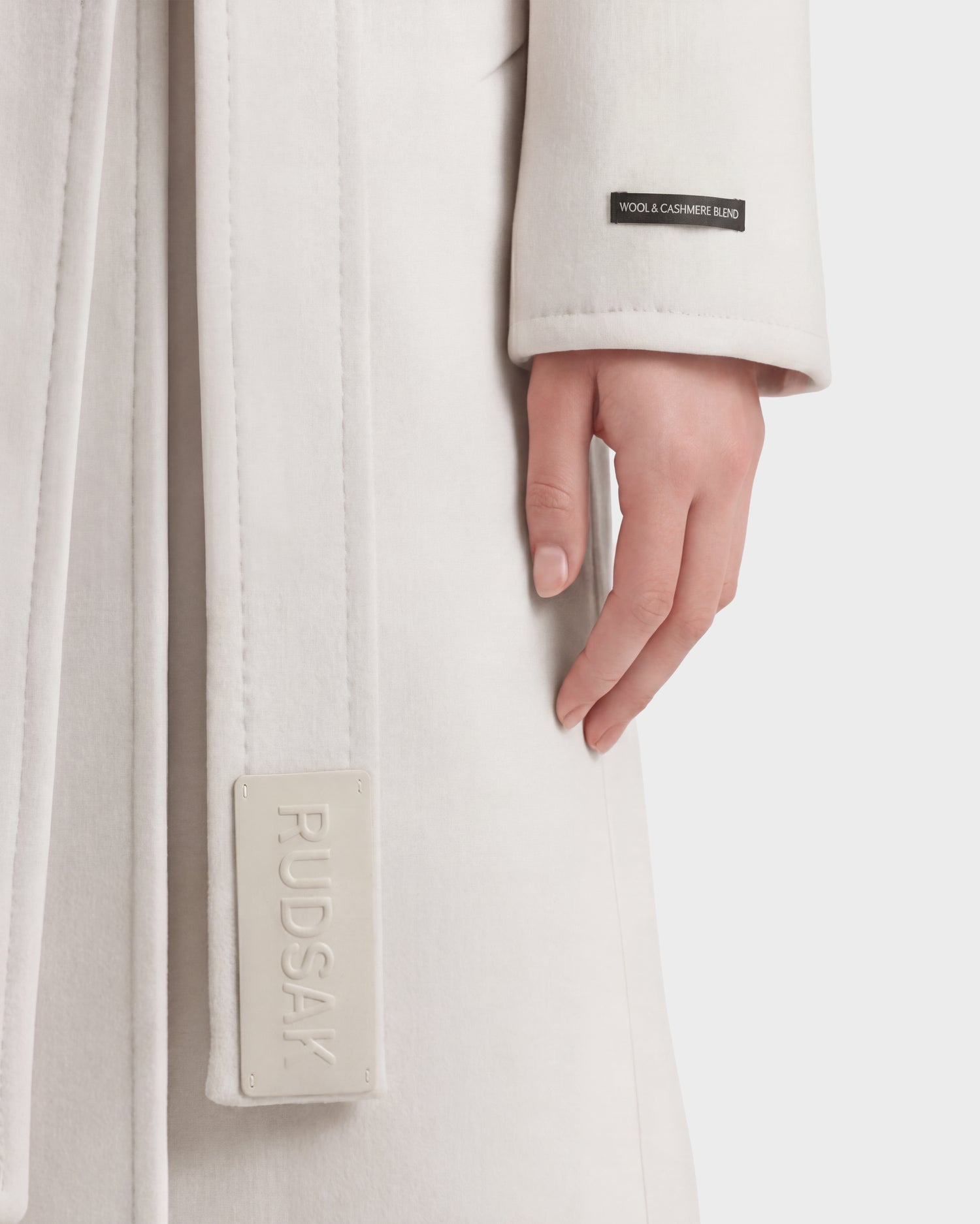 A close-up of a person wearing the KOURT W - 8124702 Vanilla coat by Rudsak, a luxurious light-colored piece made from a wool and cashmere blend. The sleeve displays a branded tag, while the person's hand rests at their side.