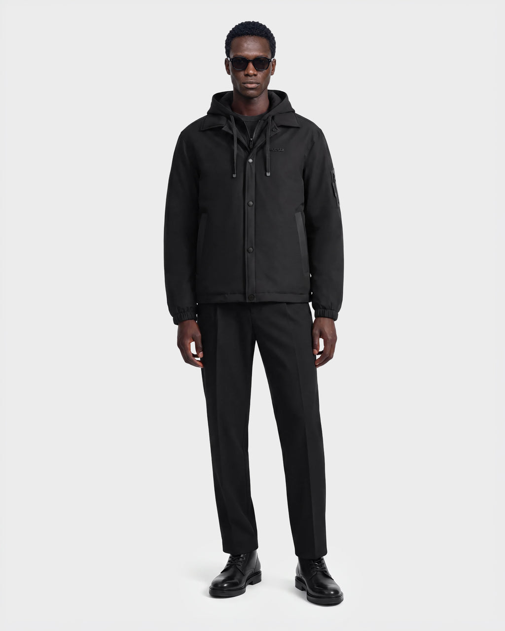 A man wearing sunglasses and the Rudsak LUCA - 6125009 Black weather-resistant hooded men’s jacket, along with black pants and black shoes, stands facing forward against a plain white background.