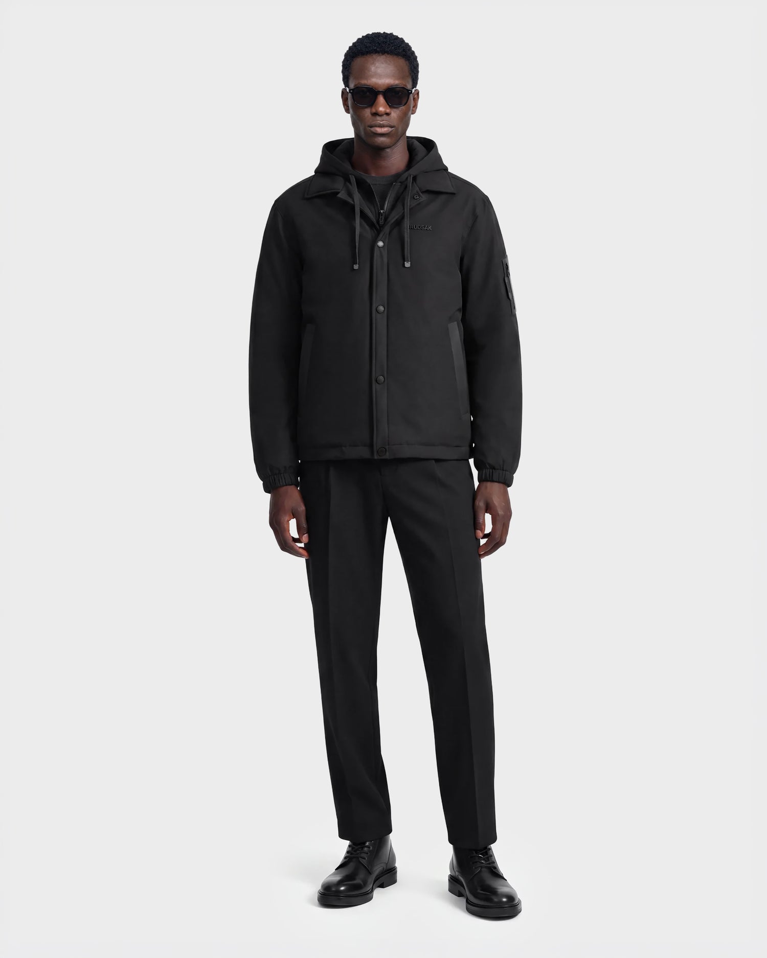 A man wearing sunglasses and the Rudsak LUCA - 6125009 Black weather-resistant hooded men’s jacket, along with black pants and black shoes, stands facing forward against a plain white background.