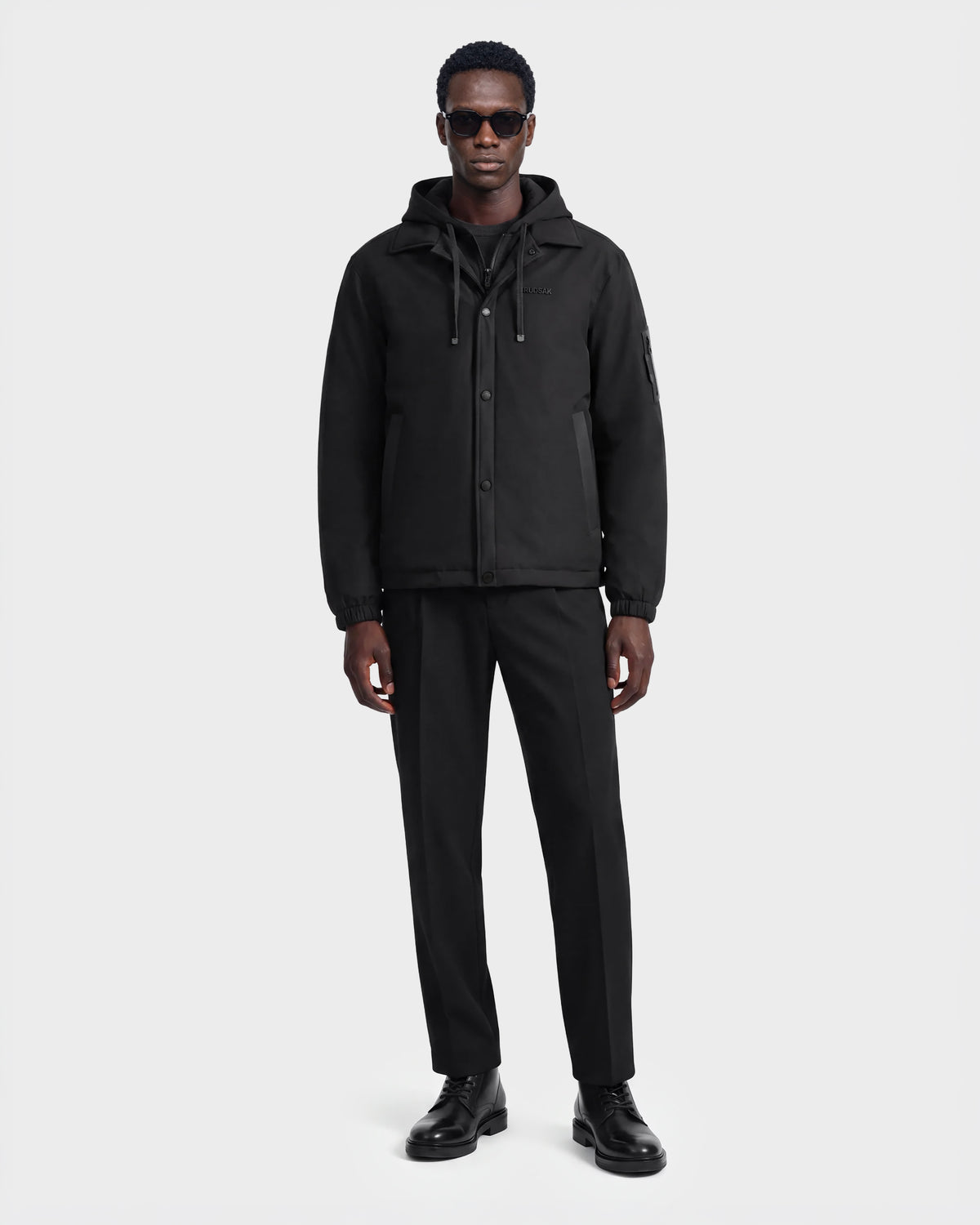 A man wearing sunglasses and the Rudsak LUCA - 6125009 Black weather-resistant hooded men’s jacket, along with black pants and black shoes, stands facing forward against a plain white background.