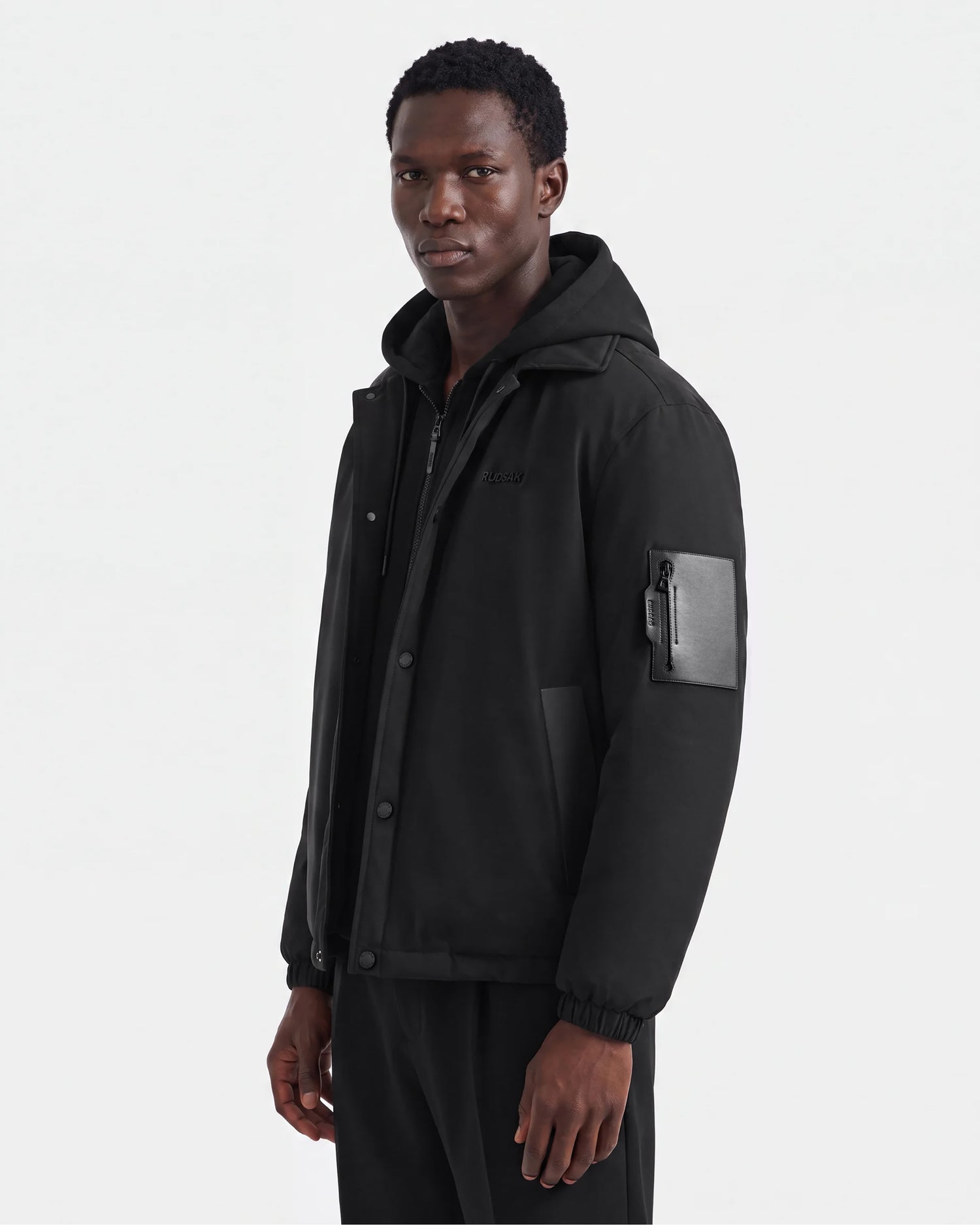 A man wearing the Rudsak LUCA - 6125009 Black men's jacket, featuring weather-resistant fabric and a zippered pocket on the left sleeve, stands against a plain white background, looking slightly to the side.