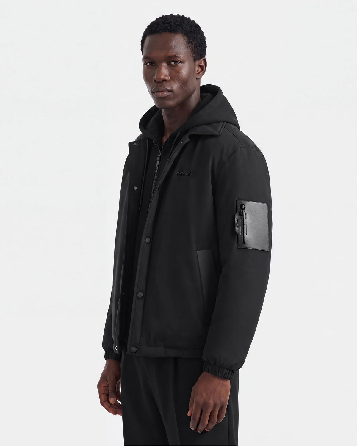 A man wearing the Rudsak LUCA - 6125009 Black men's jacket, featuring weather-resistant fabric and a zippered pocket on the left sleeve, stands against a plain white background, looking slightly to the side.