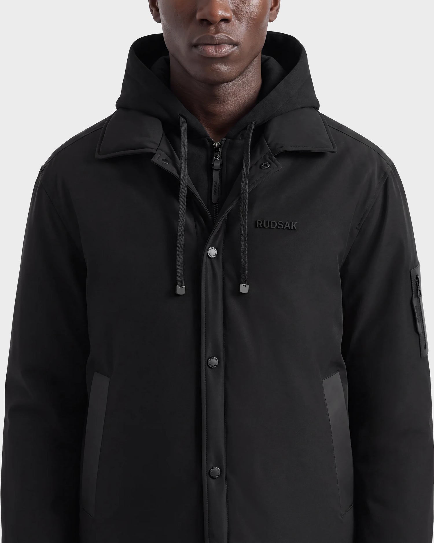 A man stands against a plain background, shown from shoulders to mid-thigh, wearing the LUCA - 6125009 Black insulated men’s jacket by Rudsak, featuring lightweight construction and RUDSAK text on the chest.
