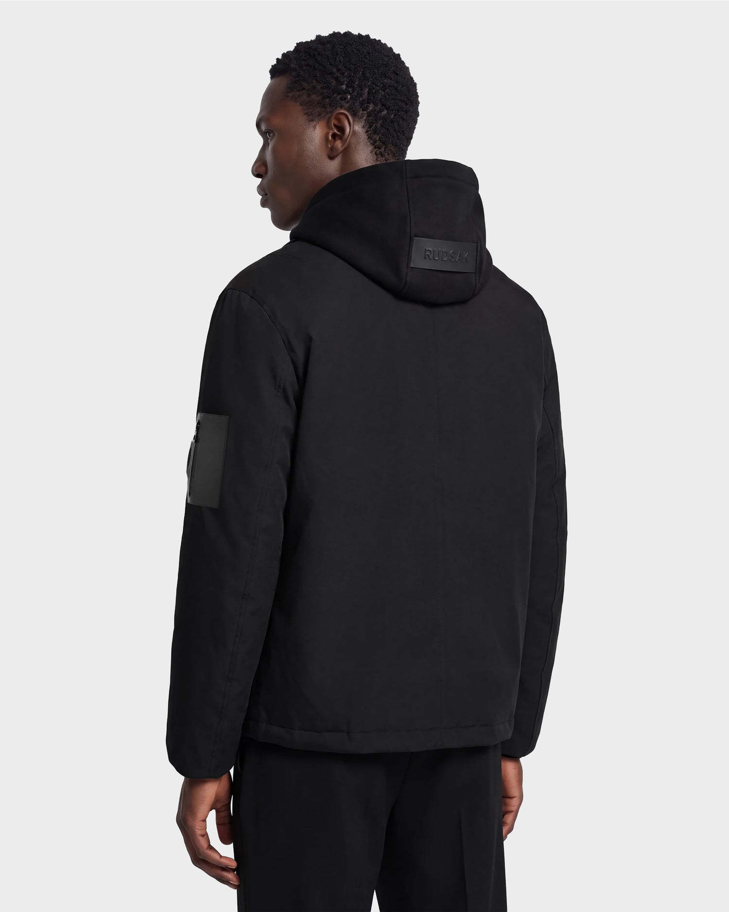 A person stands facing away, dressed in the Rudsak LUCA - 6125009 Black weather-resistant men’s hooded jacket featuring a patch on the left sleeve, paired with black pants, set against a plain light gray background.