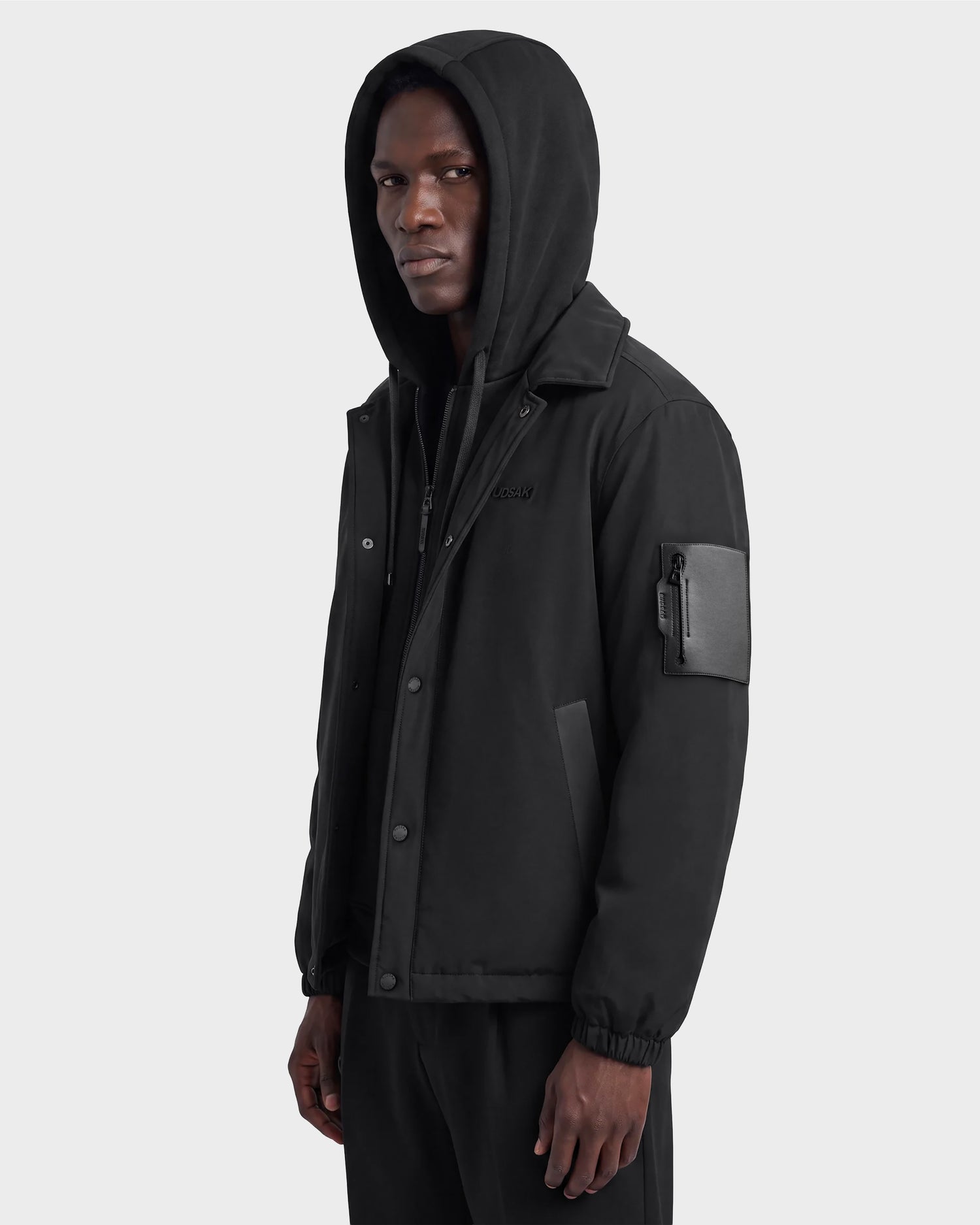 A man wearing the Rudsak LUCA - 6125009 black lightweight insulated men’s jacket, featuring a hood and a zippered sleeve pocket, stands facing slightly left against a plain light background.