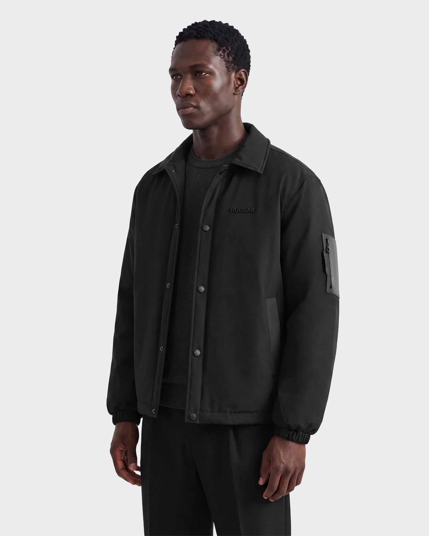 A man wearing the Rudsak LUCA - 6125009 Black weather-resistant jacket and black pants stands against a plain light gray background, looking off to the side with a neutral expression.