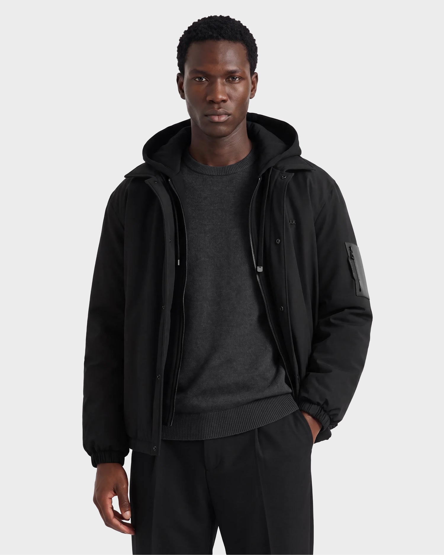 A person stands facing forward against a plain light gray background, wearing the Rudsak LUCA - 6125009 Black weather-resistant men’s jacket layered over a dark sweater and black pants, with one hand in their pocket.