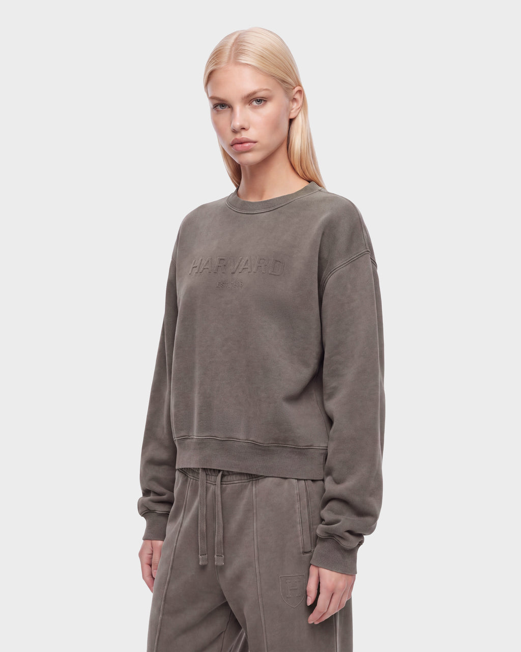 A person with straight blonde hair wears the Rudsak LEXYE HV - H526900 Taupe set, featuring a loose sweatshirt and matching sweatpants in a faded olive green shade, standing against a plain light background and looking directly at the camera.