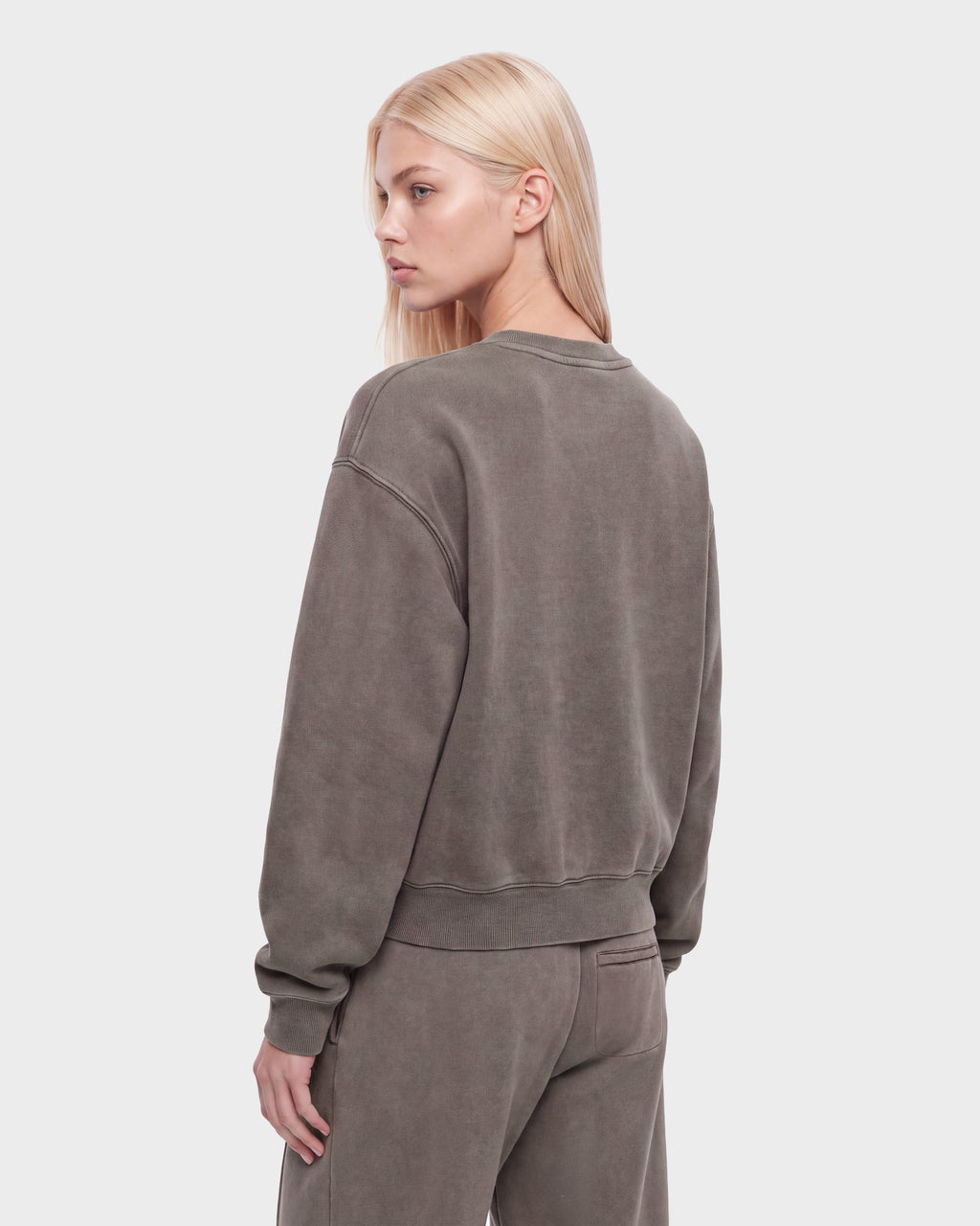 A person with long blonde hair wears the Rudsak LEXYE HV - H526900 Taupe oversized sweatshirt and matching pants, standing with their back to the camera and head turned slightly to the side.