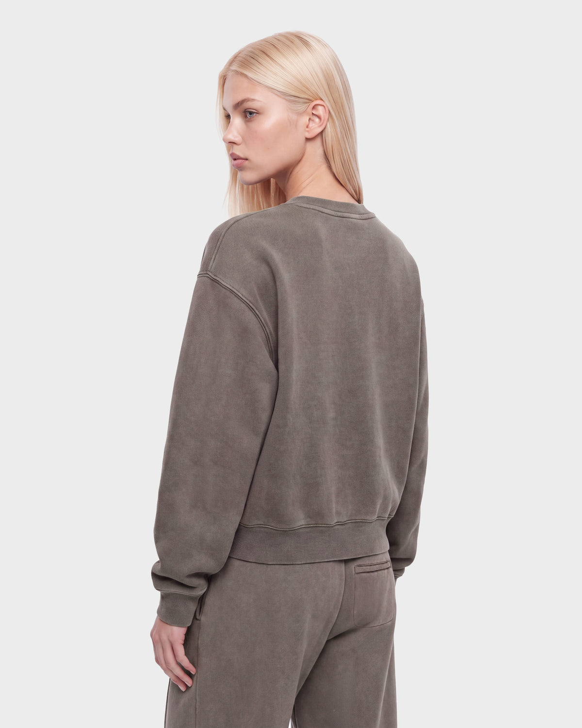 A person with long blonde hair wears the Rudsak LEXYE HV - H526900 Taupe oversized sweatshirt and matching pants, standing with their back to the camera and head turned slightly to the side.