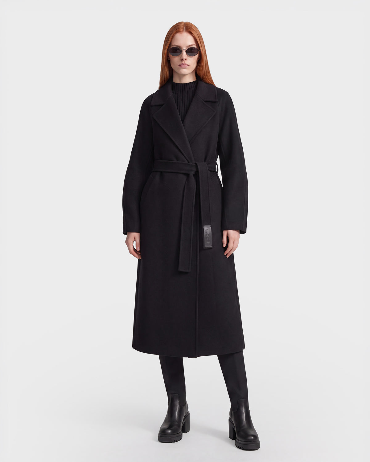 A person with long straight hair wears dark sunglasses, a long black Rudsak LILIA - 8125634 coat belted at the waist, black pants, and black boots, standing against a plain white background.