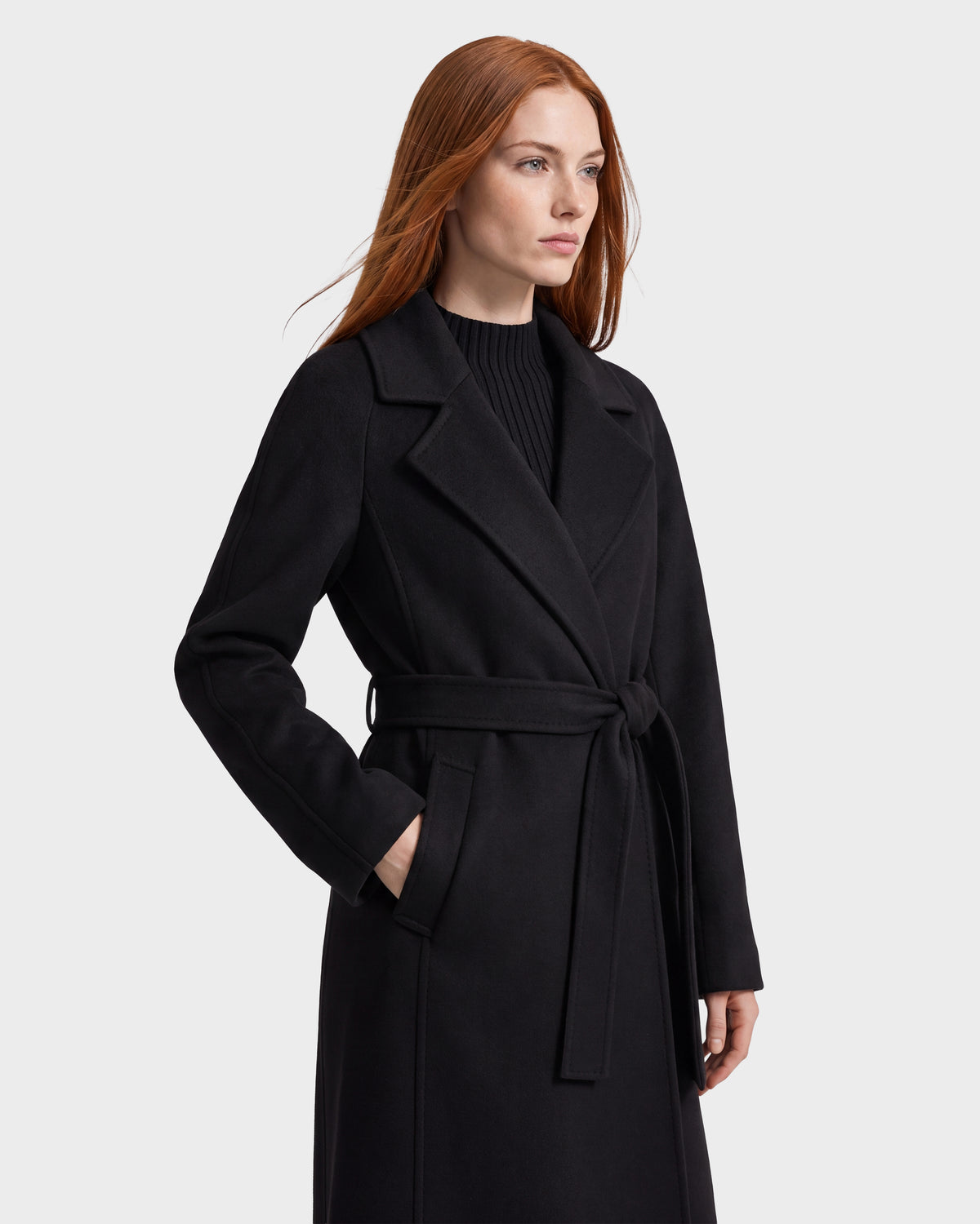A woman with long red hair wears the Rudsak LILIA - 8125634 Black coat, featuring a belted waist and shawl collar, layered over a black sweater and standing with one hand in her pocket against a plain light background.