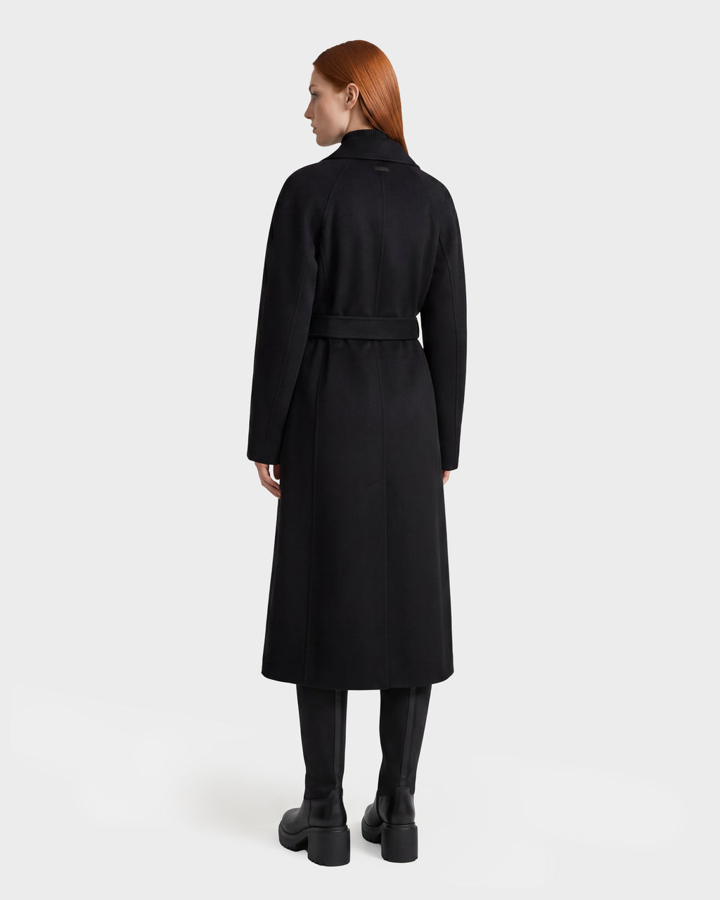 A woman with straight red hair is seen from behind, standing against a plain white background, wearing the Rudsak LILIA - 8125634 Black women’s long wrap coat belted at the waist, paired with black knee-high boots.