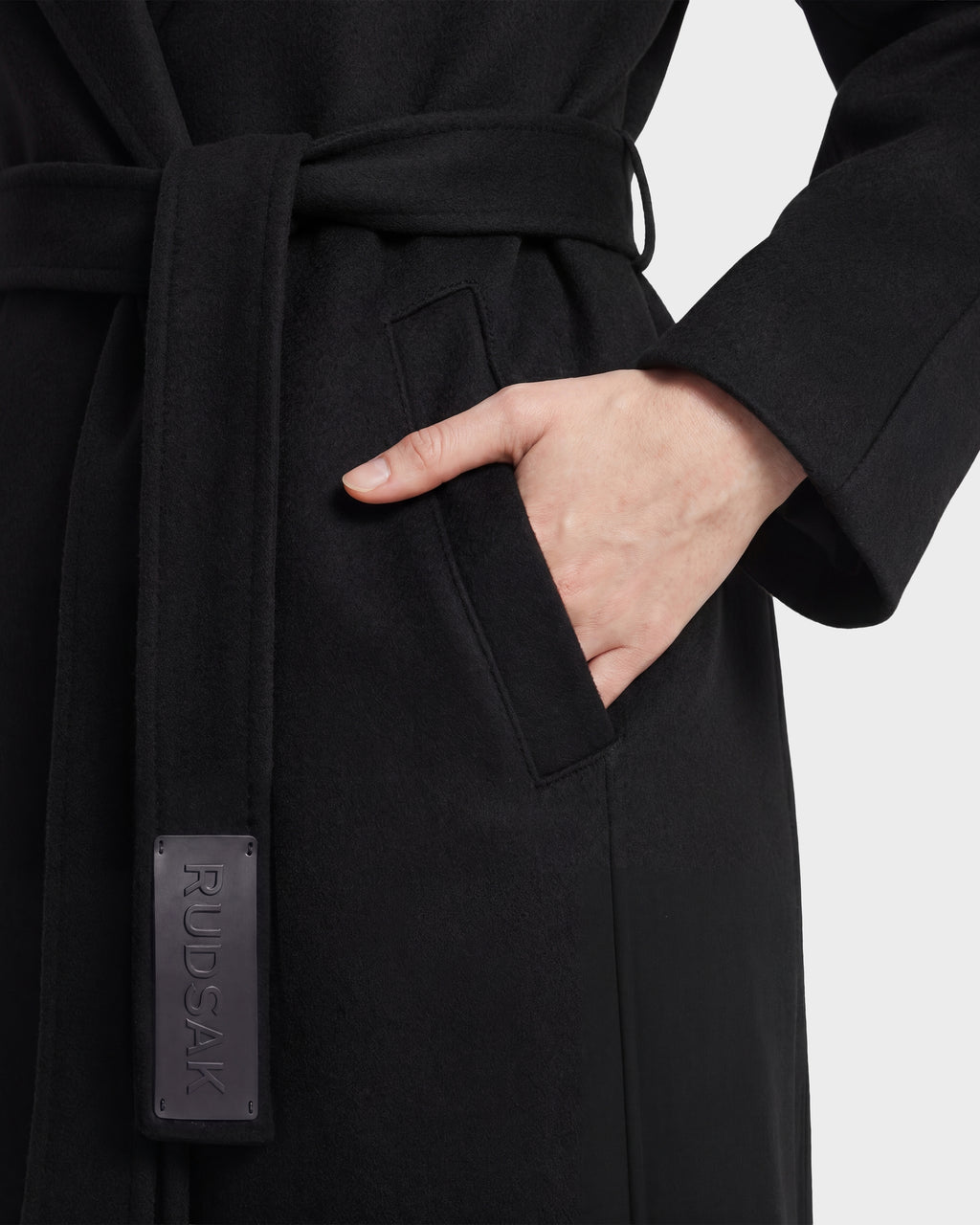 A person stands against a plain background with their hand in the pocket of a black Rudsak LILIA - 8125634 women’s wrap coat featuring a belt and a visible RUDSAK label.