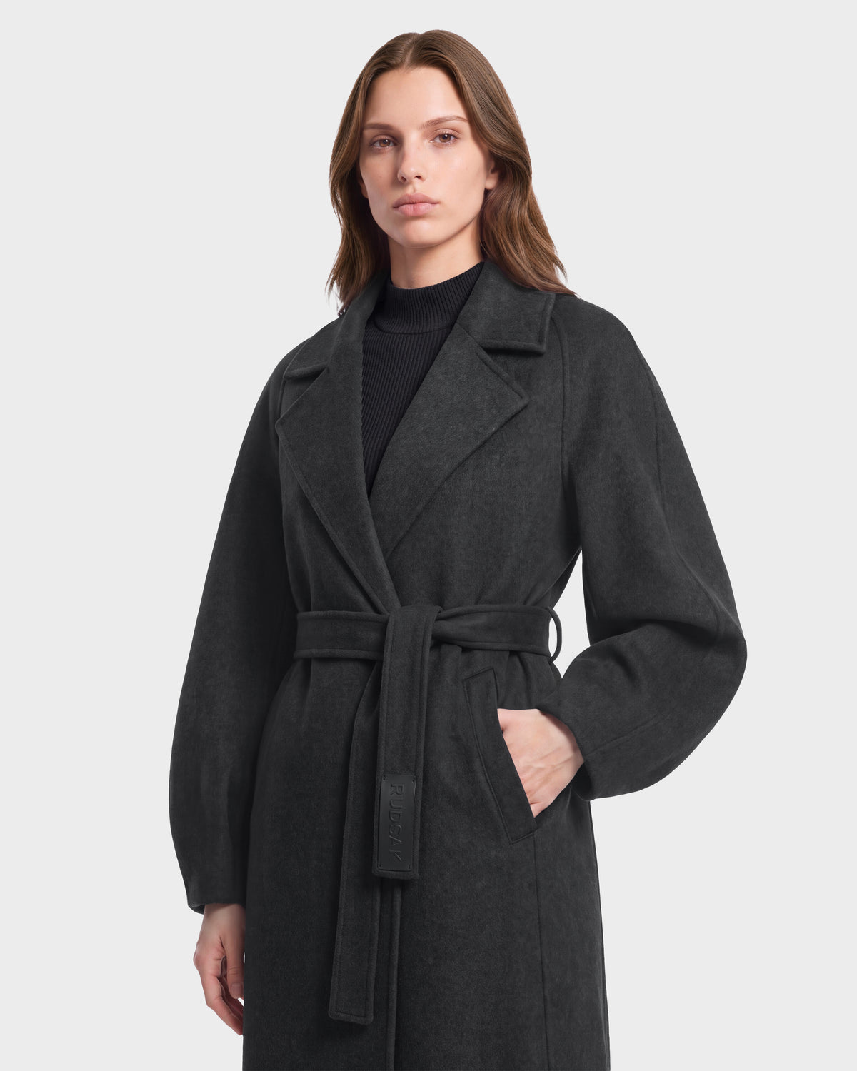 A person wearing the Rudsak LILIA - 8125634 Heather Charcoal coat with wide lapels and a black top stands against a plain light gray background, one hand in the coat pocket.