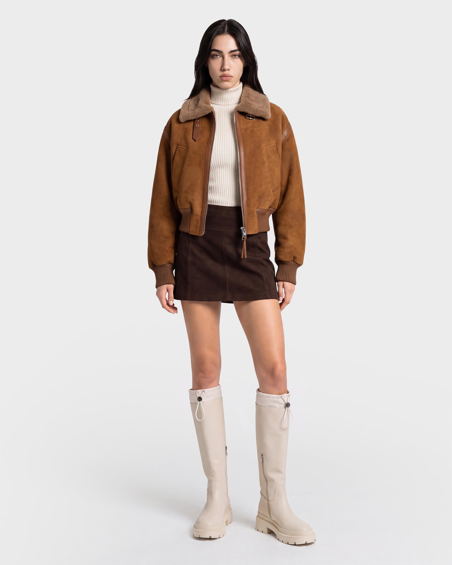 A person wearing the Rudsak LIZZIE - 8126787 Tan suede jacket, a cream turtleneck sweater, a dark brown mini skirt, and knee-high cream boots stands against a plain white background.