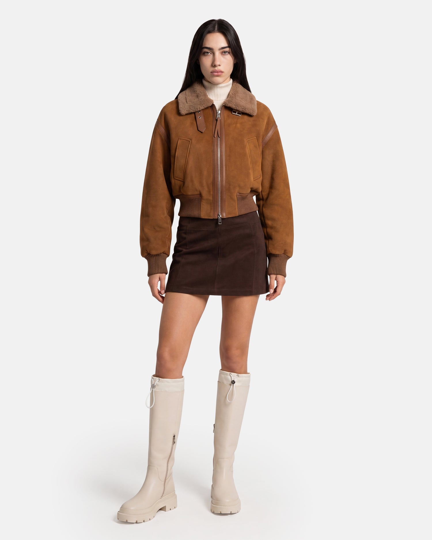 A woman stands against a plain white background wearing the Rudsak LIZZIE - 8126787 Tan shearling jacket, paired with a dark mini skirt and knee-high cream boots.