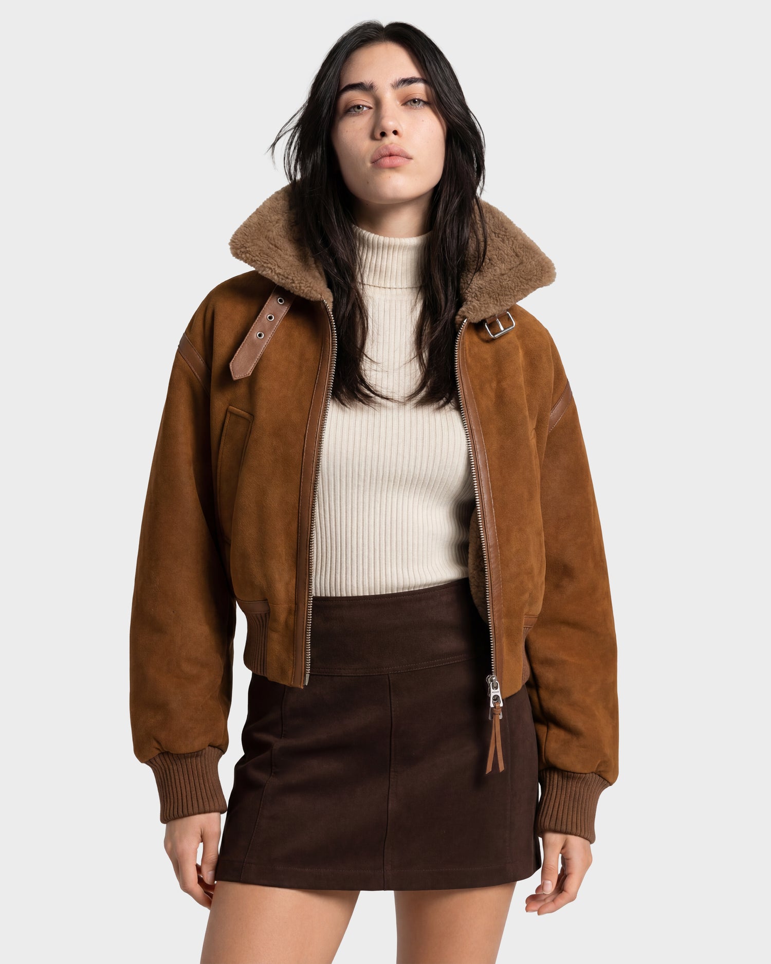 A woman stands against a plain light gray background, looking at the camera with a neutral expression, wearing the Rudsak LIZZIE - 8126787 Tan suede jacket with a fur collar, paired with a cream turtleneck sweater and a dark brown skirt.