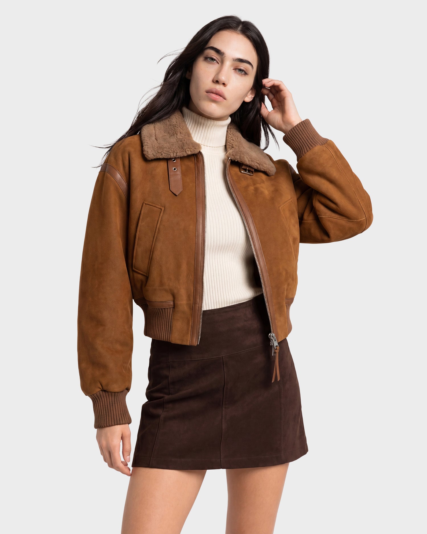 A woman wearing the Rudsak LIZZIE - 8126787 Tan shearling-collar jacket, paired with a beige turtleneck sweater and brown mini skirt, poses with one hand touching her hair against a plain background.