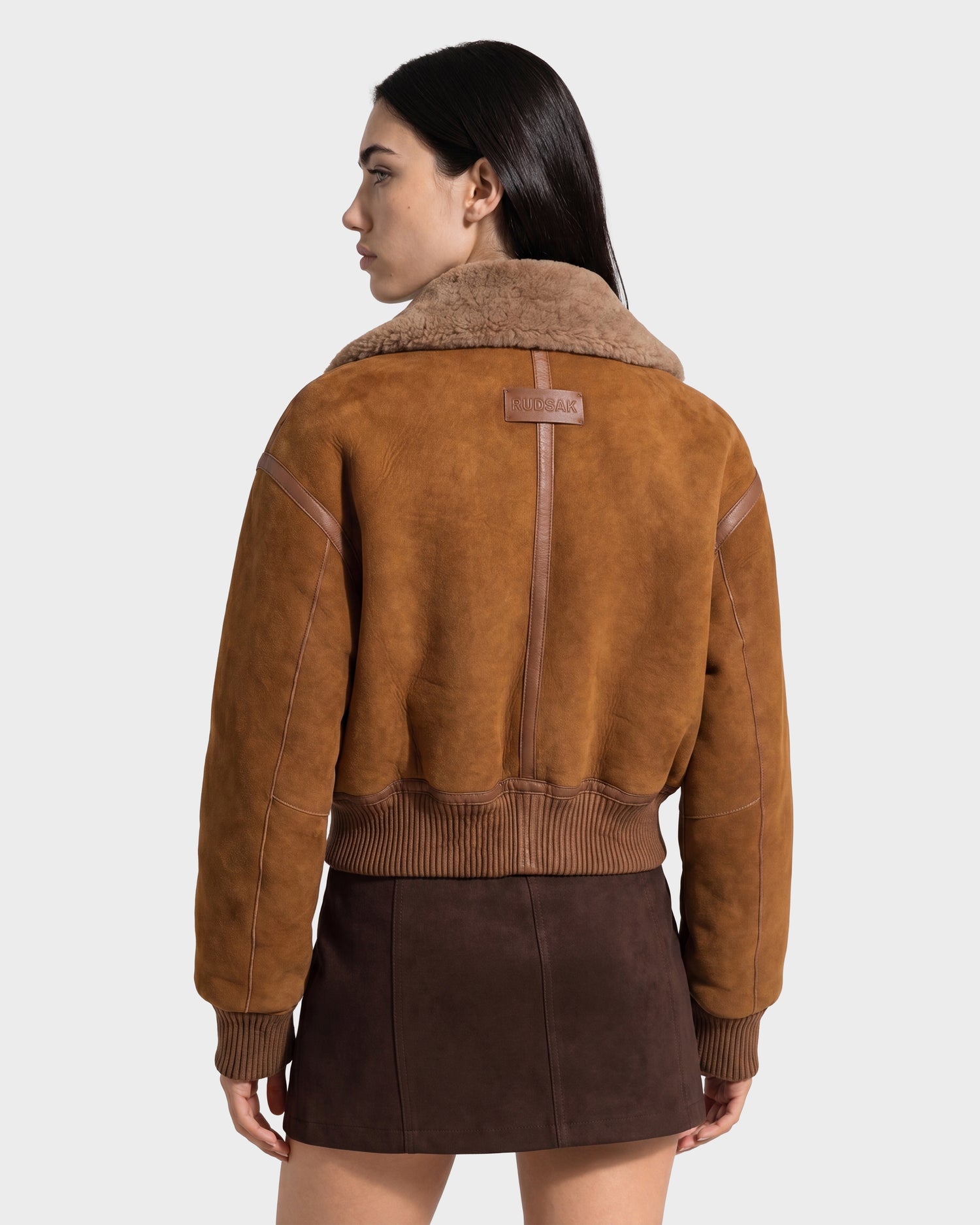 A woman with long dark hair wears the Rudsak LIZZIE - 8126787 Tan shearling bomber jacket and a dark brown mini skirt, standing and looking to the side against a plain background.