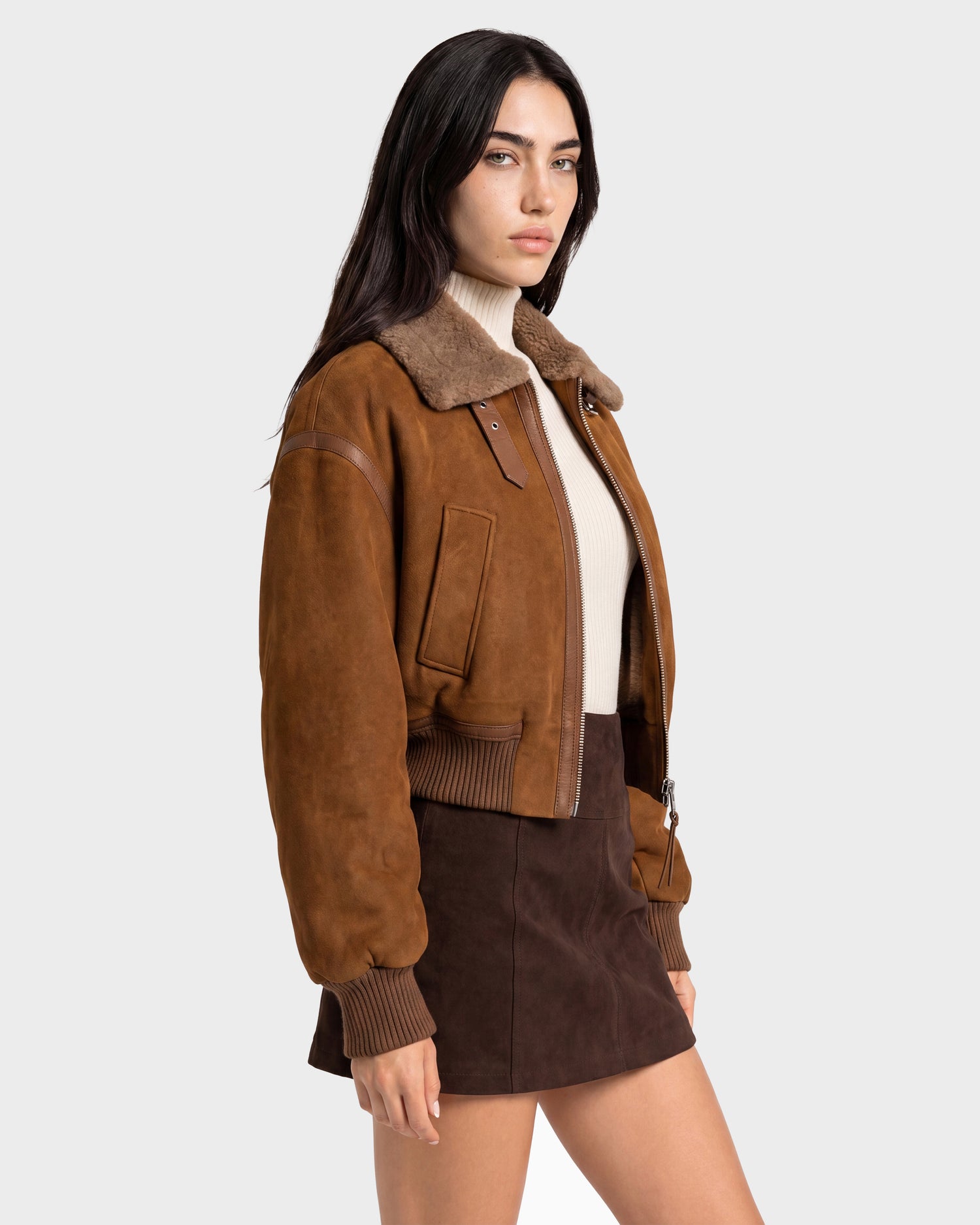 A woman wearing the Rudsak LIZZIE - 8126787 Tan shearling jacket over a beige turtleneck and brown mini skirt stands against a plain light gray background.